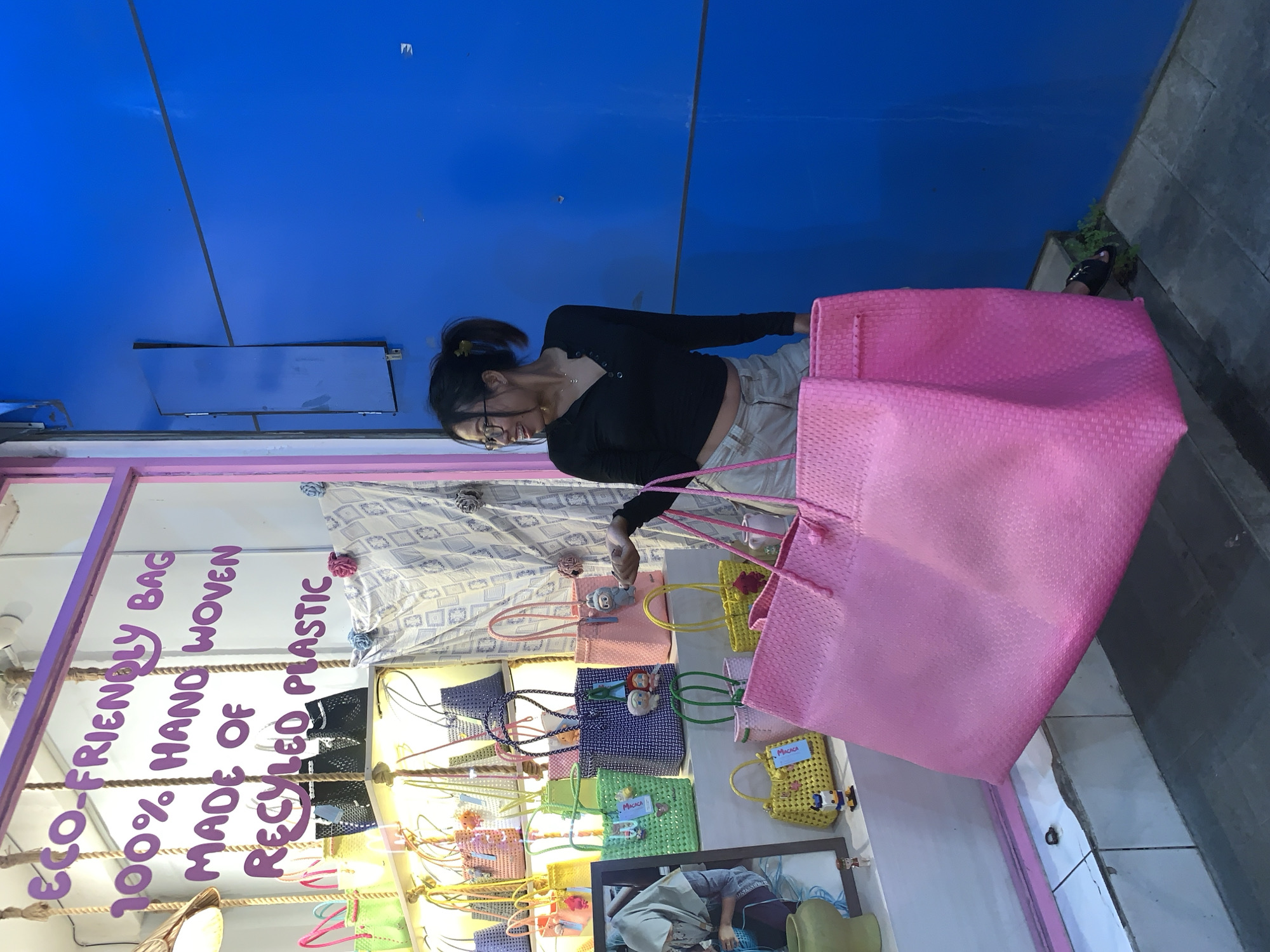 A young woman stands in front of a shop window in Ubud District, Indonesia. She is wearing a black top and light-colored pants, and she has her hair pulled back in a bun. She is looking at a display of colorful woven bags made from recycled plastic. The shop window has a sign that reads "Eco-friendly bag, 100% hand woven, made of recycled plastic." The bags are arranged on shelves, and there are several different colors and styles. The woman is holding a large pink bag. The background of the image is a blue wall and a sidewalk.