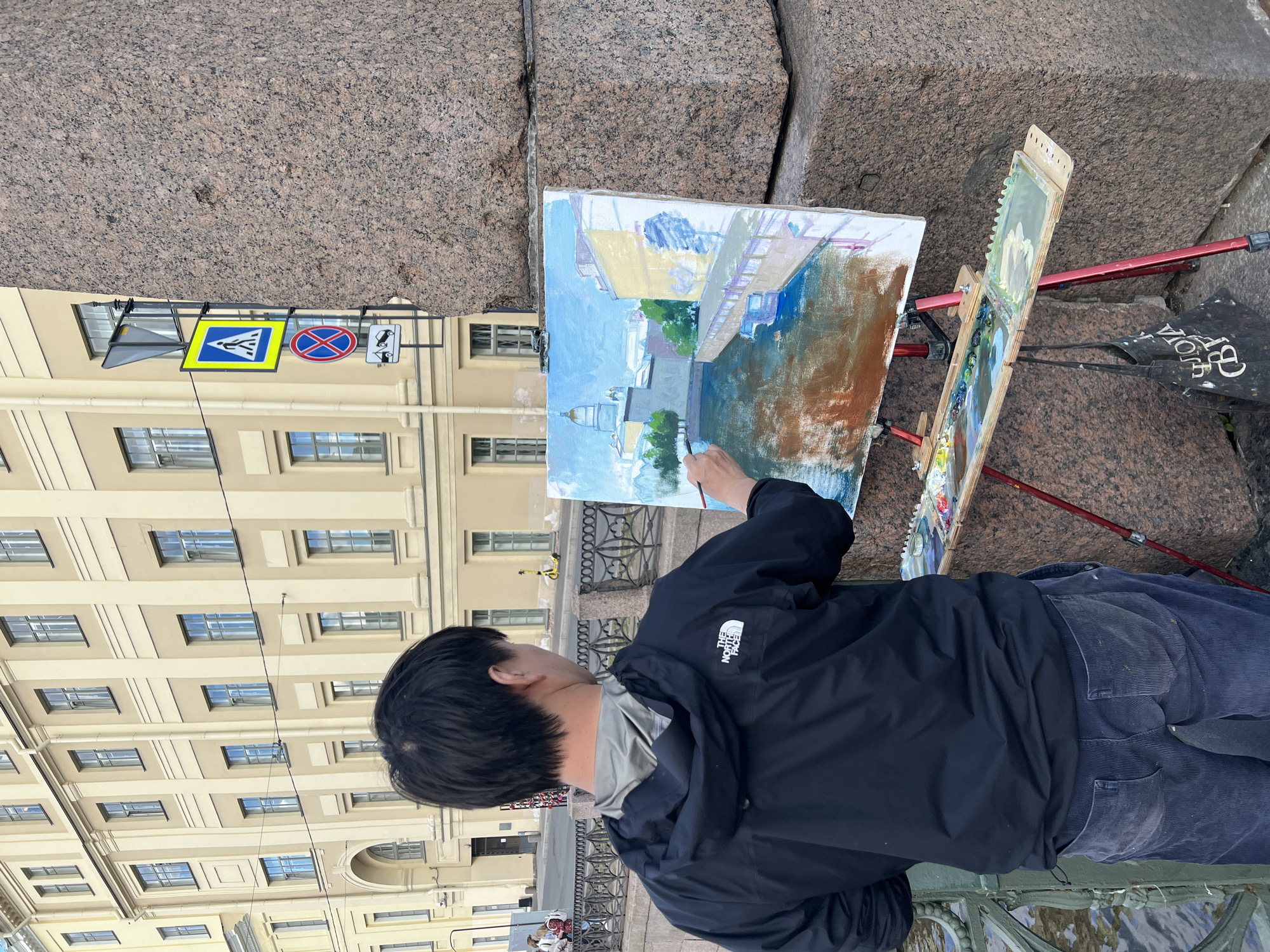 The image shows a person painting a landscape outdoors in Saint Petersburg, Russia. The artist, viewed from behind, is wearing a black jacket and dark pants, and is focused on a canvas set up on a red easel. The painting on the easel appears to be a cityscape with buildings and water. Beside the main canvas, a smaller painting is also visible on the easel, showing a few figures.
The background features a large granite building with rows of windows, indicative of classical architecture often found in Saint Petersburg. Traffic signs, including a pedestrian crossing sign and a "no entry" sign, are visible in the upper left corner, suggesting the painting is taking place near a street. The lighting suggests it is daytime, with overcast skies or diffused light, which is common in Saint Petersburg, especially outside of peak summer. The overall scene conveys a sense of artistic focus amidst an urban environment.
There is visible text on the artist's jacket, reading "THE NORTH FACE". Additionally, there is text on a black bag resting near the easel, though it is partially obscured and difficult to fully decipher. The granite facade and the architectural style of the building strongly suggest a location within the historic center of Saint Petersburg.