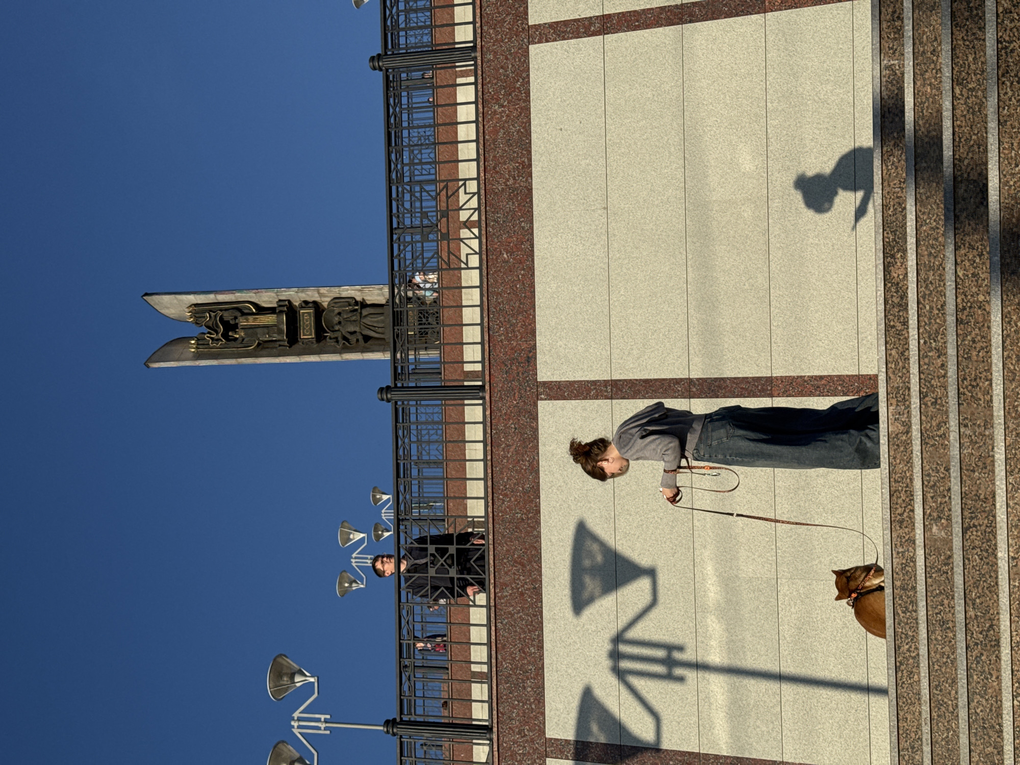 The image displays a multi-level outdoor urban scene under a clear blue sky, depicting the Monument to the Friendship of Peoples (also known as the Friendship Stele) in Izhevsk, Russia, along with surrounding architectural elements and individuals.
At the upper level, a tall, split monument with a central golden relief bearing the letters "CССР" (USSR) and various figures is prominently featured on the right side. Several street lamps with conical diffusers are visible against the sky on the left and center. A black metal railing with intricate patterns spans the upper level. Behind this railing, a male figure wearing a dark coat and glasses stands, looking towards the left. Other individuals are partially visible further back on this upper platform, obscured by the railing. The wall supporting the upper level features an upper band of reddish-brown speckled granite and a lower section of light beige textured panels.
On the lower level, a female figure with her hair tied in a bun is standing on the uppermost step of a wide granite staircase. She is wearing a grey hooded sweatshirt and loose-fitting blue jeans and holds a dog leash in her right hand. A reddish-brown dog, possibly a Shiba Inu or similar breed, sits calmly on a lower step to her left, facing away from the camera, wearing a harness and attached to the leash. The steps are made of speckled reddish-brown and grey granite with metallic accents. Elongated shadows of the upper-level street lamps are cast onto the beige wall, creating geometric patterns. The shadow of the female figure, with a distinct bun and appearing to hold a device in her hand, is cast onto the beige wall to the right. The lighting indicates a low sun angle, suggesting either morning or late afternoon.