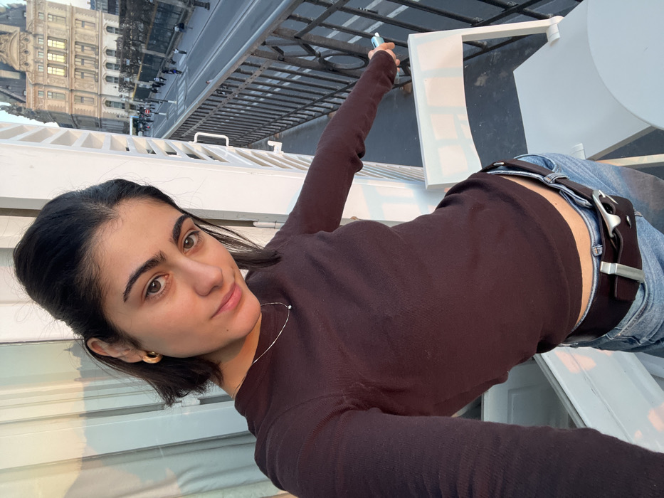 A young woman is depicted in a selfie photograph, standing on a balcony overlooking Rue de Rivoli in Paris, France, directly opposite a portion of the Louvre Palace. The woman, appearing to be in her late teens or early twenties, has dark, shoulder-length hair partially pulled back, brown eyes, and prominent eyebrows, exhibiting a faint smile. She wears a fitted, long-sleeved dark brown top, light-wash blue jeans, a dark brown leather belt with a silver-toned buckle, a delicate silver-colored necklace with a small pendant, and gold-toned hoop earrings. Her left arm extends, holding a small, light blue cylindrical object resting on the black wrought-iron balcony railing. A light-colored outdoor chair is visible to her right. A white louvered shutter is partially open to her left. Below, pedestrian and vehicle traffic is observed on the street. Warm, late-day sunlight illuminates the scene, casting areas into shadow.