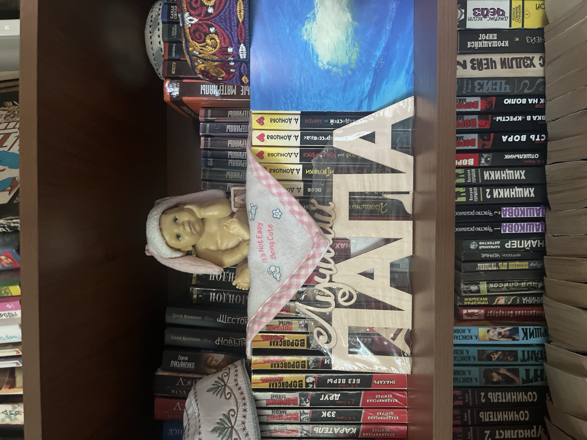 The image depicts a bookshelf filled with various books. On the top shelf, a doll resembling a baby wearing a pink bunny hat is placed between two decorative hats, one white and one embroidered with red and gold patterns. A pink and white checkered cloth with a printed message "It's Not Easy Being Cute" is laid out in front of the doll. Wooden letters spelling out "Лучший ПАПА" (Best Dad) are displayed on the shelf. The middle shelf also contains books, with some of their spines visible. The bottom shelf is entirely filled with books, some with colorful covers. The overall environment suggests a home interior, specifically a room with a bookshelf. There is no information or visual evidence in the image to suggest any connection to the city of Vladimirovka, Russia, or any specific geographic location.