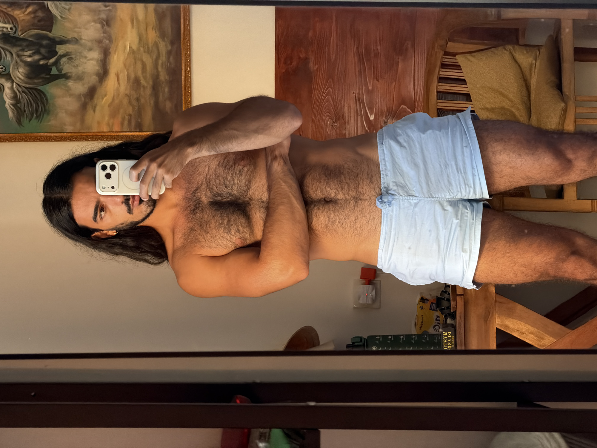 A shirtless, hirsute man with long dark hair holds a white phone up to his face, seemingly taking a mirror selfie. He is wearing light blue shorts. Behind him, the background includes a painting with horses, a wooden wall or furniture piece, and a chair with a beige cushion. The location seems to be a domestic setting, potentially in North Kuta, Indonesia. Other objects in the reflection include a book with green text, a pack of instant noodles, and a wooden structure, possibly a shelf or a table.