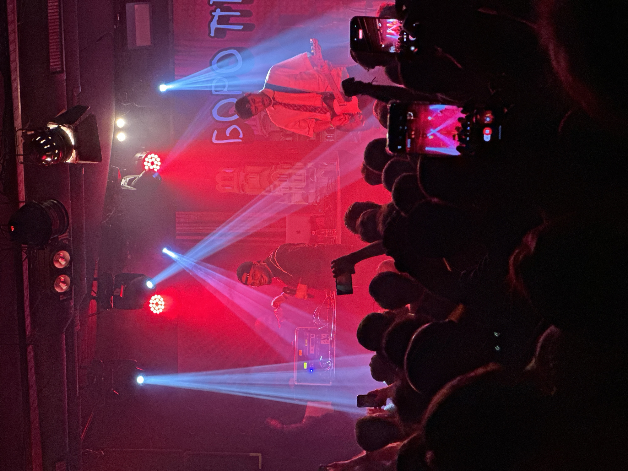 The image depicts a live musical performance, likely a concert. Two individuals are visible on stage, illuminated by a combination of red and blue stage lights. One person is dressed in a white shirt and tie while playing a guitar. The other person is wearing darker clothing and appears to be singing or rapping into a microphone. A crowd of people is visible in the foreground, many holding up their mobile phones to record the performance. Based on the signage visible, it's possible this is happening in Sofia, Bulgaria. There is also concert lighting and staging visible.