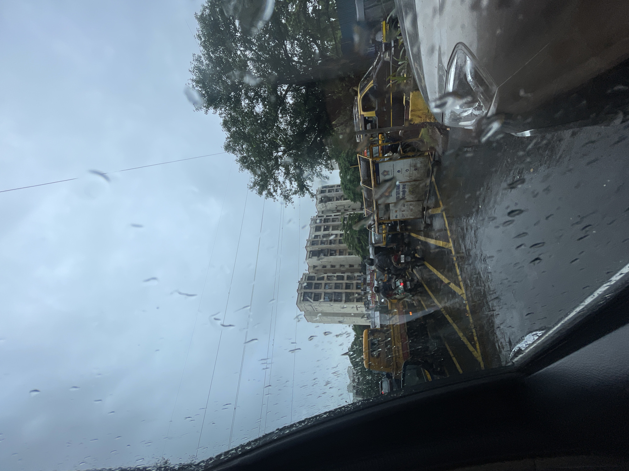 The image captures a scene of city traffic in Mumbai, India, during a downpour. The perspective is from inside a vehicle, looking out through a rain-streaked windshield. In the background, a multi-story residential building stands under a cloudy, overcast sky, indicative of monsoon weather. A large, leafy tree is visible on the right side of the frame.
The road is wet and reflects the surrounding environment, with yellow road markings clearly visible. A line of vehicles, including auto-rickshaws and motorcycles, are stuck in traffic. Several people are riding on the motorcycles, some wearing helmets. A yellow truck is also part of the traffic jam. The overall atmosphere suggests a typical day during Mumbai's monsoon season, characterized by heavy rain and traffic congestion.