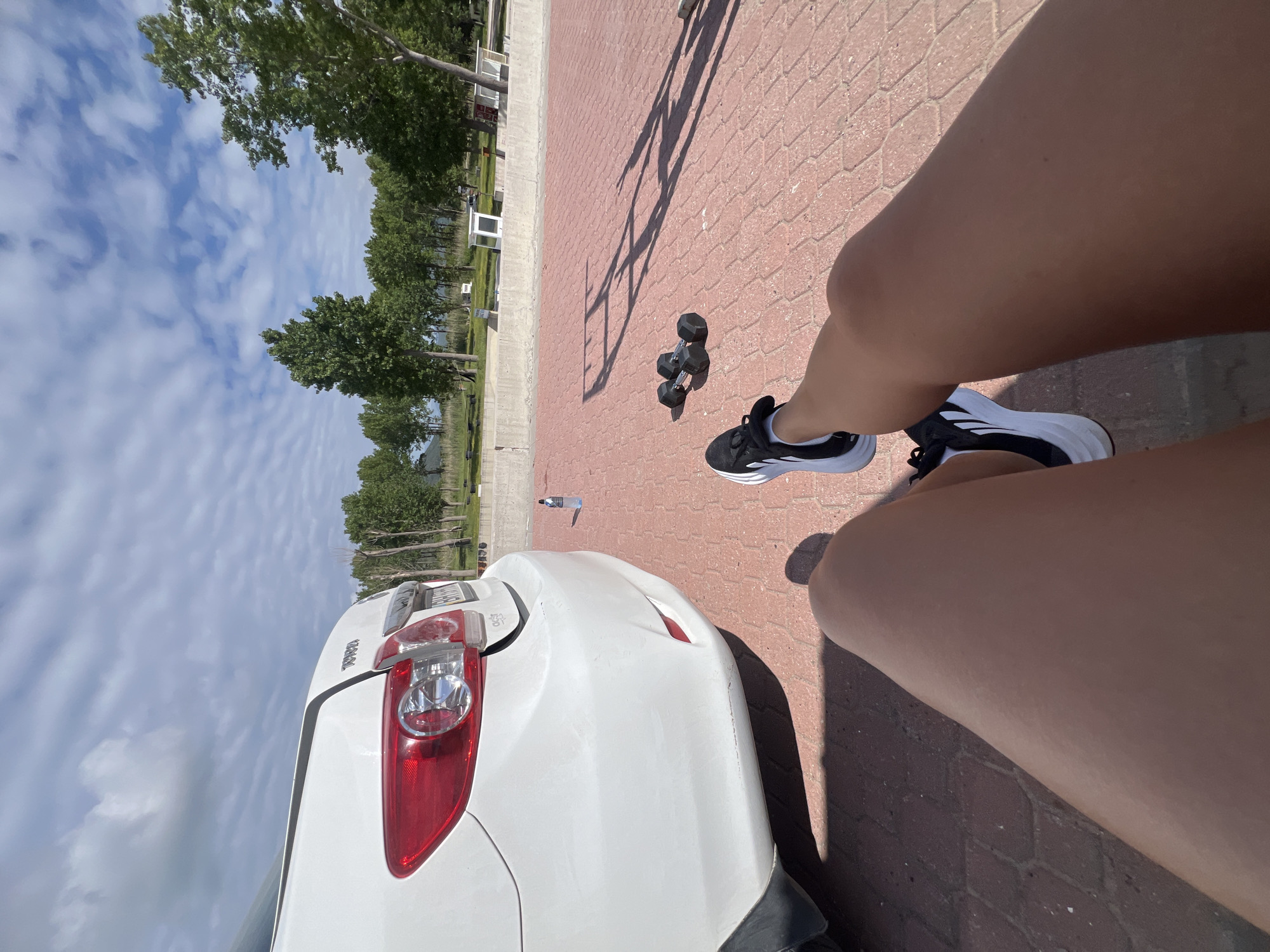 A high-angle, rotated perspective captures a sunny outdoor scene on a brick-paved area near what appears to be a waterfront or park. In the foreground are the legs of a person wearing black and white athletic shoes. Next to the legs are a pair of black dumbbells. Nearby is a white car, the rear of which is partially in view. The background shows trees, a blue sky with scattered clouds, and hints of buildings. A bottle is also visible. The specific location is in the city of Prijevor, Montenegro.