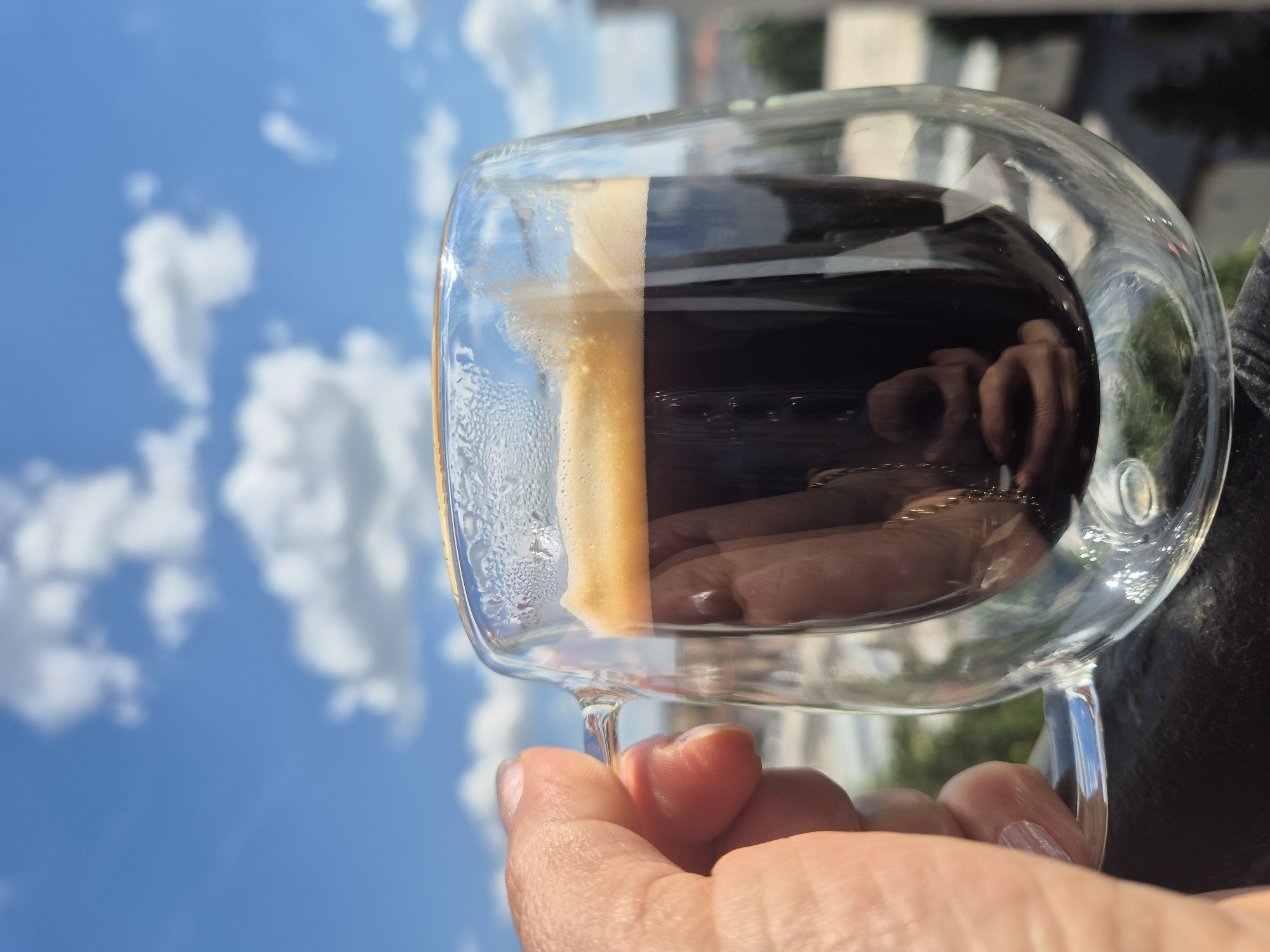 A close-up shot shows a hand holding a double-walled glass mug filled with dark espresso coffee, topped with a frothy crema. The mug is held against a bright blue sky dotted with fluffy white clouds. In the blurred background, a cityscape with buildings and trees is visible, suggesting an outdoor setting in an urban area. The lighting indicates a sunny day.