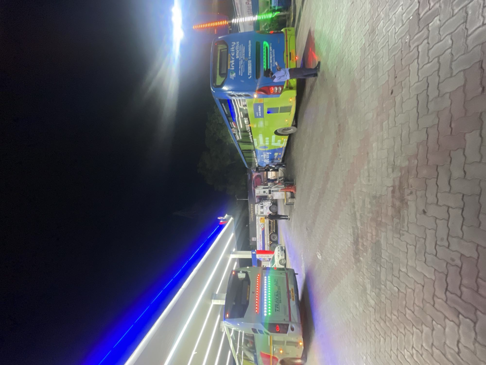 At a brightly lit gas station in Kajheri, India, a fleet of buses and trucks are parked under a covered canopy illuminated by white and blue LED lights. The ground is paved with interlocking bricks.
On the left, a white and green bus with "TRAVEL AP" written on its rear is parked. The bus's taillights are illuminated with a red and green pattern.
In the center, a white car is parked, and further back, a fuel tanker truck with "POWERES" written on it is also visible.
On the right, a blue and green IntrCity smartbus is parked. A man in a blue uniform, likely an attendant, stands near the rear of the IntrCity bus, holding a device. The bus features "IntrCity smartbus" branding and contact information. Green and orange lights are visible to the right of the bus. The scene is captured at night.