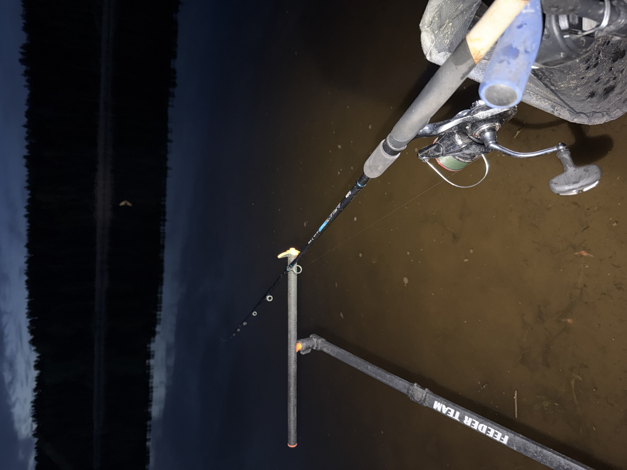 A fishing rod and reel are positioned at the edge of a body of water, likely the Kama River in Chaikovsky, Russia, given the user's location. The rod is resting on a stand labeled "FEEDER TEAM," with the line extending into the water. A fishing net is partially visible behind the reel. The background shows a dark, reflective surface suggesting either night or twilight. The overall environment appears to be a riverbank or shoreline.