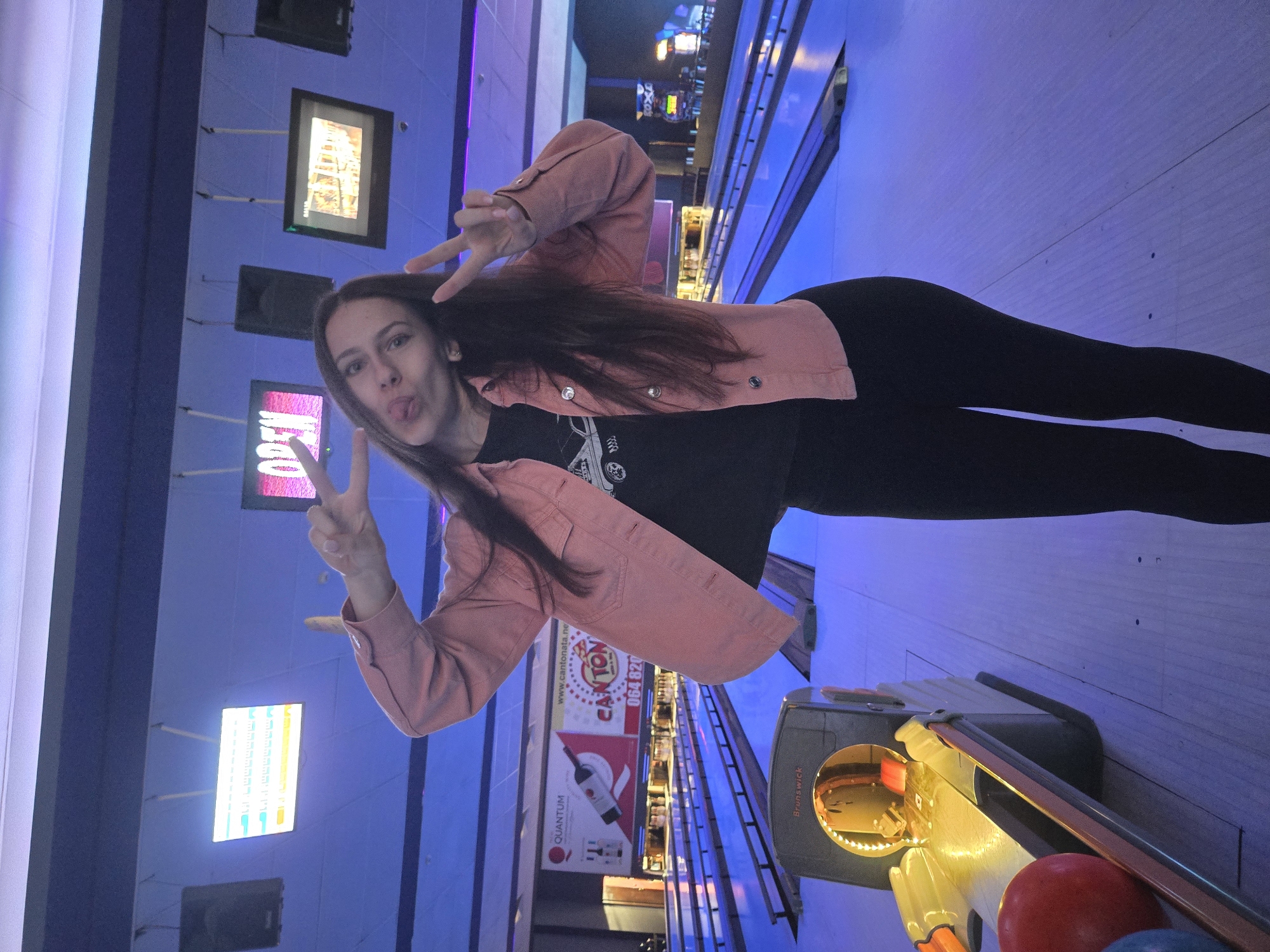 A woman stands in a bowling alley in Pleven, Bulgaria. She is wearing black leggings, a black t-shirt, and a light pink jacket. She is making a peace sign with both hands and sticking out her tongue. The bowling alley has multiple lanes with electronic scoring systems above each lane. Bowling equipment is visible near the woman. Advertisements and signs are present on the walls.