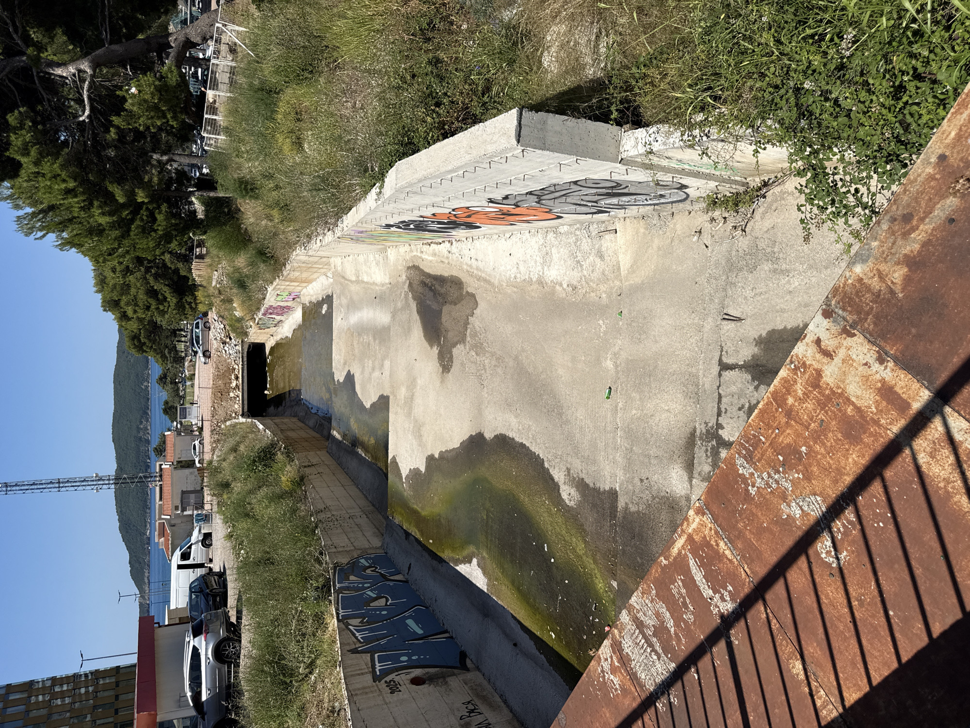 An elevated, eye-level, long shot captures a concrete drainage channel in Rafailovići, Montenegro, on a clear, sunny day. The channel is partially filled with murky, greenish water and debris. The concrete walls are covered in graffiti, with a prominent blue graffiti tag on the left wall and orange and grey graffiti on the right wall. Overgrown vegetation encroaches on the sides of the channel. In the background, beyond a line of trees, are the blue waters of the Adriatic Sea, a hilly coastline, and buildings. Cars are visible parked near a building on the left side of the frame. A rusty metal railing is in the foreground, casting a shadow across the scene.