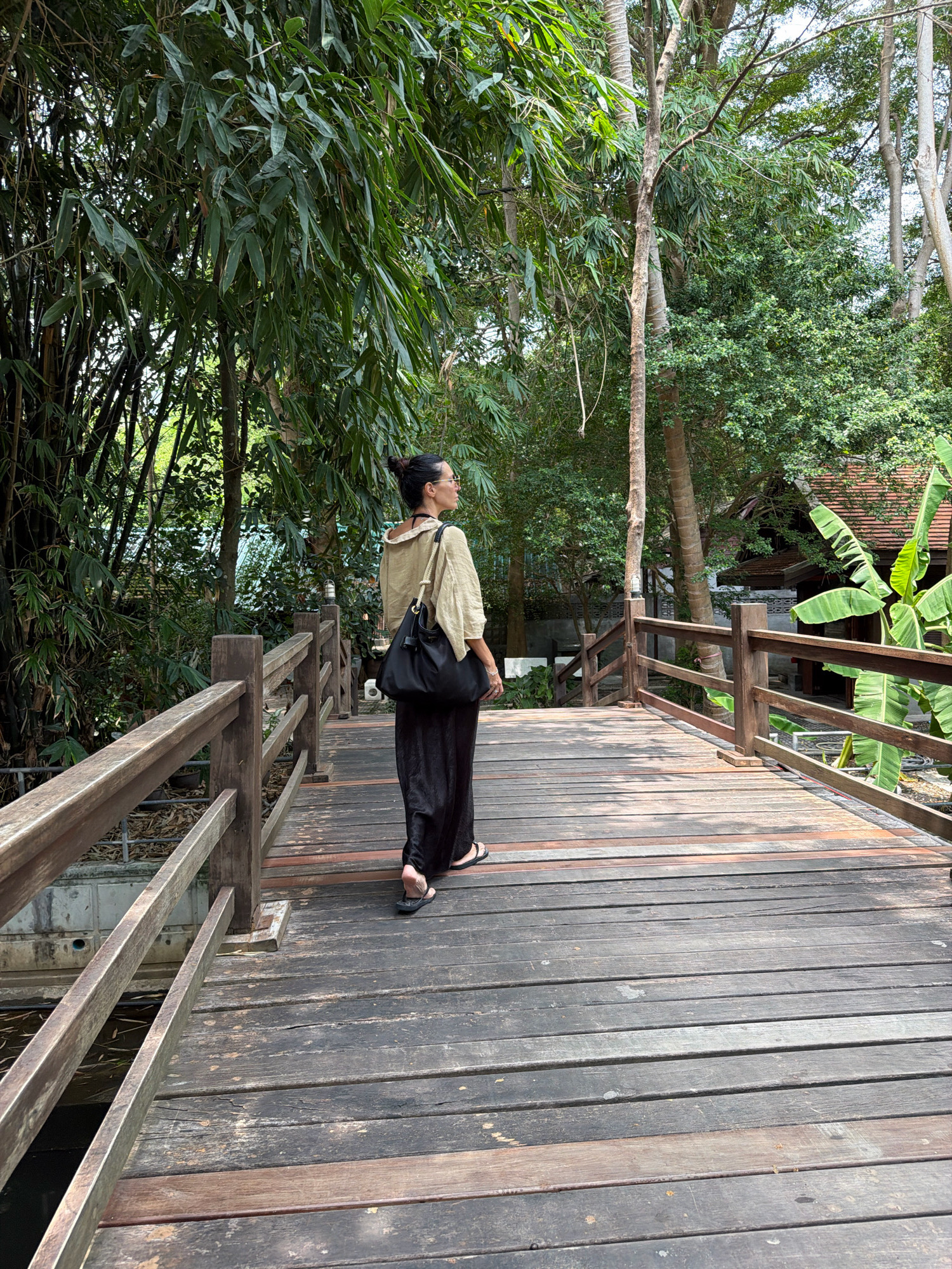 A lone woman walks on a wooden bridge in a lush, green environment. She is seen from behind, wearing a light-colored, loose-fitting top and dark, wide-legged pants. She has a dark shoulder bag and her hair is pulled back in a bun. The bridge is constructed of weathered wooden planks and has wooden railings. Surrounding the bridge is dense vegetation, including bamboo and palm trees, suggesting a tropical setting, likely in Ko Samui District, Thailand. In the background, a traditional Thai-style building with a tiled roof is partially visible. The scene appears to be a pathway through a garden or park.