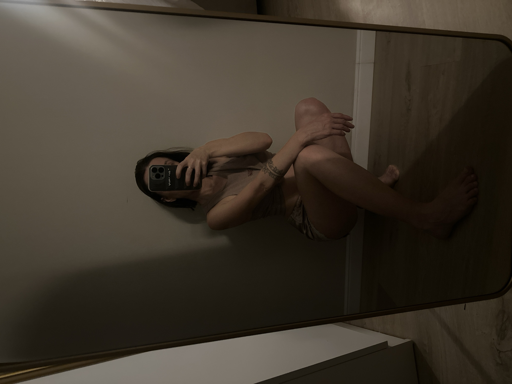 A woman is taking a selfie in a mirror. She is wearing a beige top and shorts and sitting on the floor. The background shows a wall and a wooden floor. The location is impossible to determine from the image.