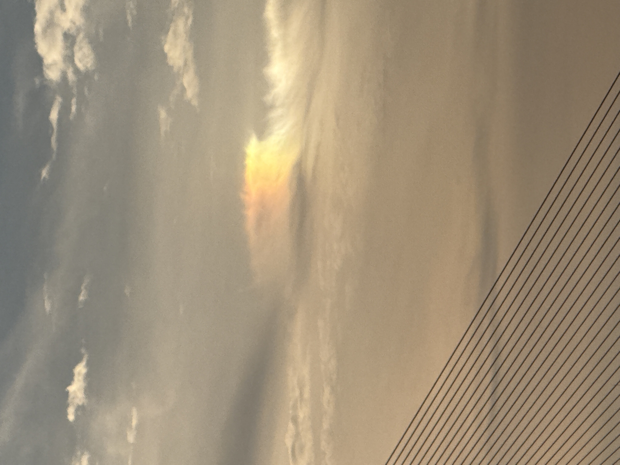 This is a photograph of a sky with clouds and a portion of a bridge. The sky is overcast with some lighter patches and a distinct iridescent cloud formation that resembles a rainbow or sun dog. The lower right portion of the image shows the cables of a suspension bridge, indicative of the Đà Nẵng, Vietnam location, specifically the Đà Nẵng Bridge. The time of day appears to be either sunrise or sunset due to the warm, golden hues in the sky and the angle of light. The weather is cloudy, but there are breaks in the clouds allowing sunlight to refract through ice crystals, creating the colorful optical phenomenon in the sky. There is no visible text in the image.