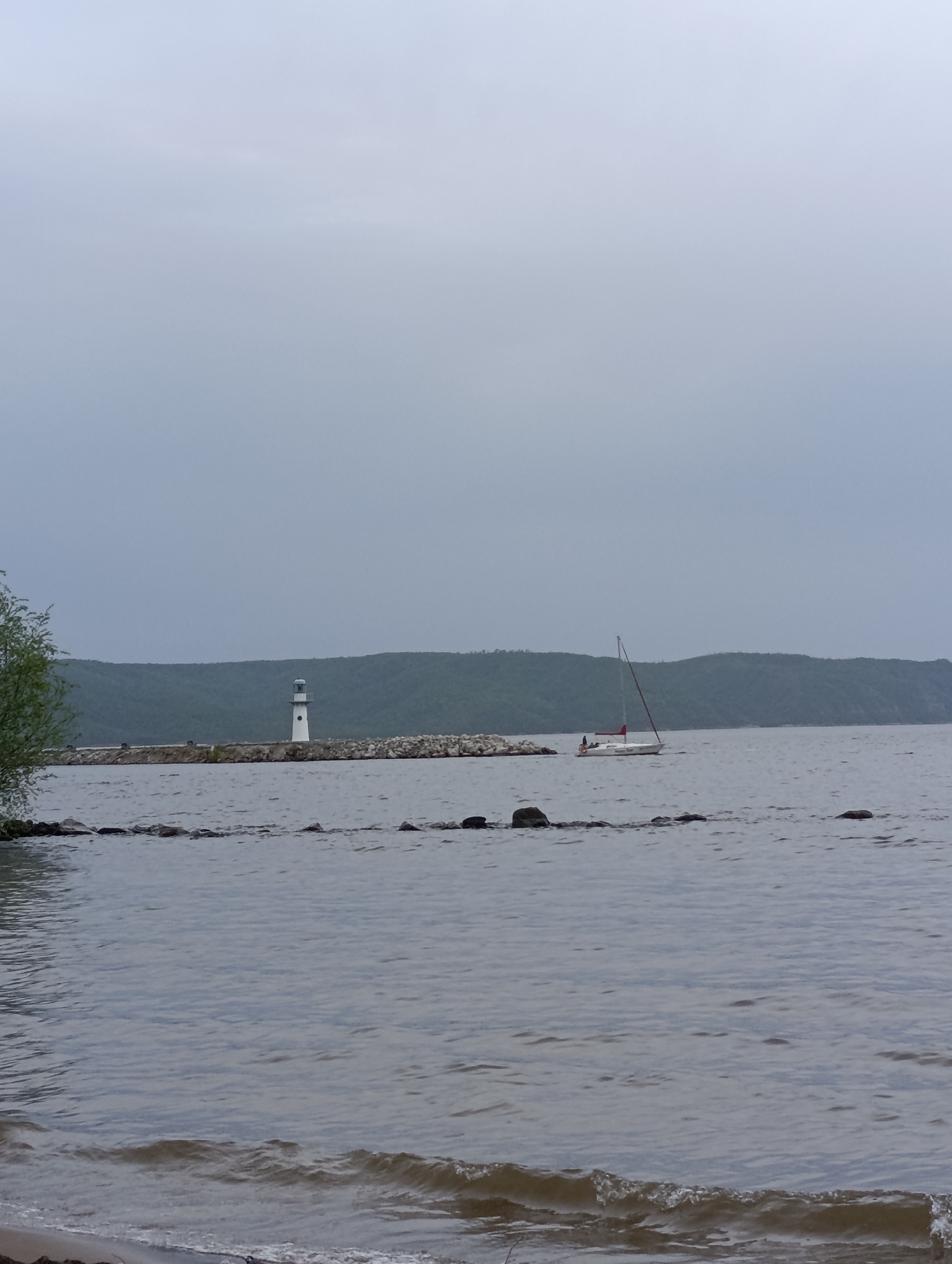 The image shows a body of water with small waves near a shoreline. A white lighthouse stands on a rock pier extending into the water. A sailboat with a red sail is visible further out in the water. A landmass with trees forms the backdrop. The sky is overcast. The location is Unknown, Unknown.