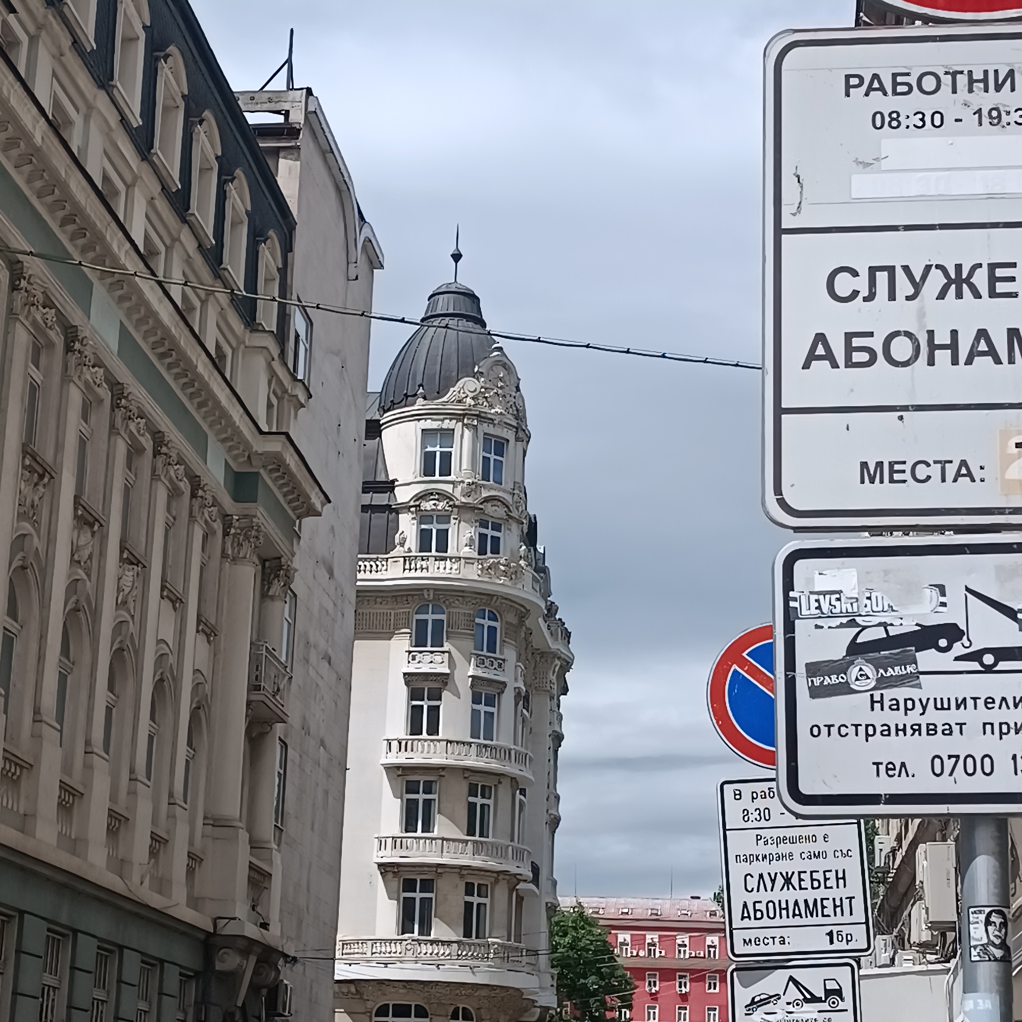 This image captures an urban street scene in Sofia, Bulgaria, characterized by historical architecture and a dense display of street signs. The foreground is dominated by a collection of white regulatory signs mounted on a pole, partially obscuring the view of buildings beyond. Utility wires crisscross the frame against an overcast, bright sky.
To the left, a tall, ornate building with classical architectural elements, such as decorative window frames, cornices, and balconies, occupies a significant portion of the frame. Its facade is a light color, possibly cream or white, with some sections having a light blue-green hue. Further back and to the right, a striking, light-colored building with a distinctive dark-domed roof and multiple balconies stands out. Its architecture is elaborate, featuring intricate carvings and decorative balustrades. In the distant background, a vibrant red building with numerous windows and a repeating roofline adds a splash of color, with a small tree visible in front of it.
No people or vehicles are visible in the shot, presenting a quiet, static street view. The time of day appears to be daytime, under a cloudy sky.
Several Cyrillic signs convey parking regulations and information:
* A large sign at the top indicates "РАБОТНИ" (Working) hours from "08:30 - 19:30", followed by "СЛУЖЕБЕН АБОНАМЕНТ" (Service/Official Subscription) and "МЕСТА:" (Places/Spots:).
* Another sign below it features an image of a tow truck, warning "Нарушители отстраняват при тел. 0700 13" (Violators are removed at phone 0700 13...). A sticker with "ПРАВО С ЛЪВЧЕ" (Right with a lion cub) is affixed to this sign.
* A smaller sign further down specifies: "В раб 8:30 -" (During working hours 8:30 -), "Разрешено е паркиране само със СЛУЖЕБЕН АБОНАМЕНТ" (Parking is permitted only with a Service/Official Subscription), and "места; 1бр." (places; 1 pc.).
* A "No stopping" or "No parking" sign (red circle with blue diagonal) is partially visible behind the parking signs.
* A sticker with "LEVS50" and another with "ЗА" (for) are also present on the signs and lamppost.