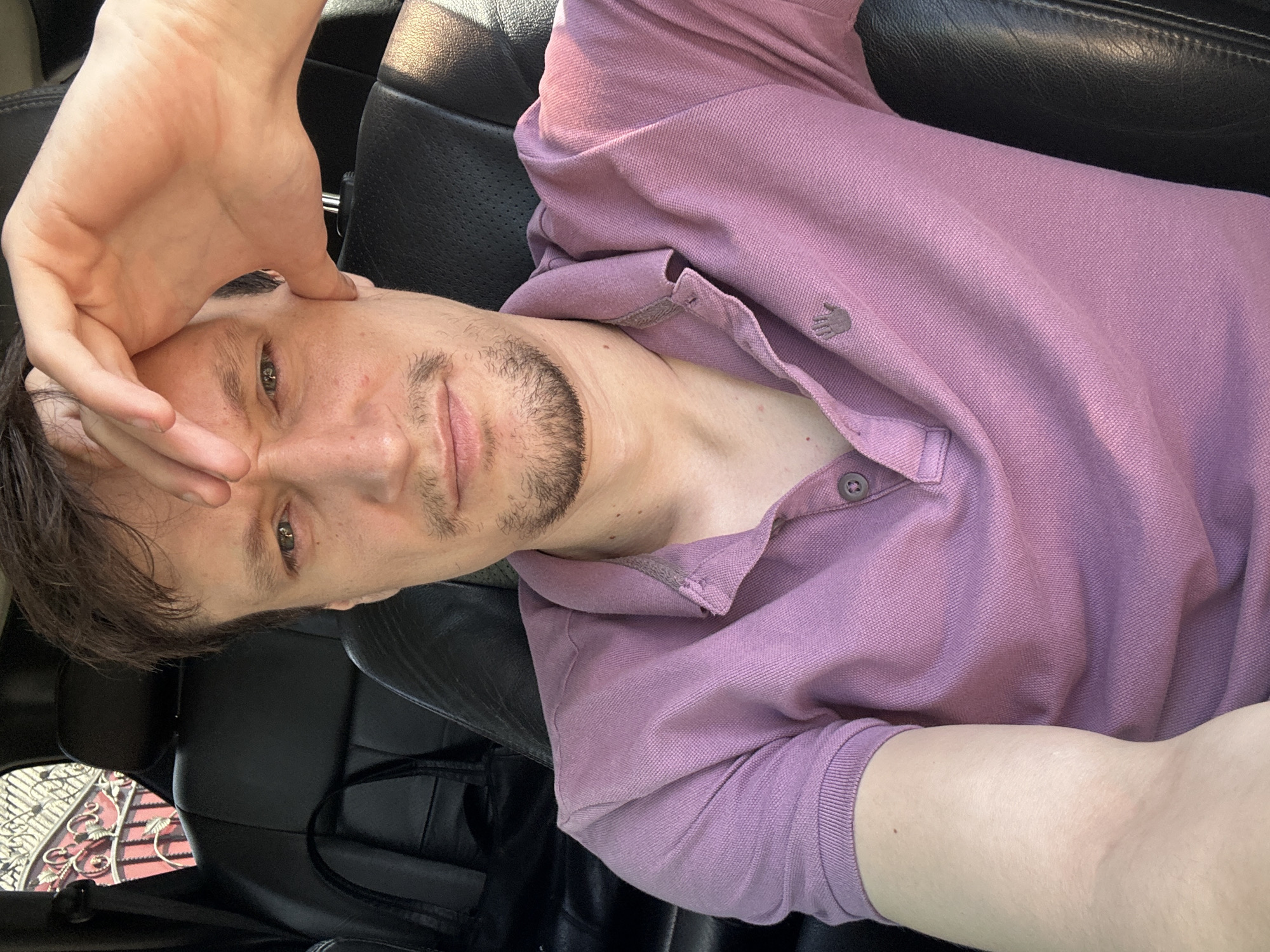 A man is sitting in the driver's seat of a vehicle, presumably parked. He is looking directly at the camera with his right hand resting on his forehead. He is wearing a light purple polo shirt with a button-up collar and a small embroidered logo on the left chest area. The interior of the car appears to be black leather. The background visible through the rear window shows a decorative metal fence with red elements and some foliage. There is no indication of the specific location within the city of Makiv, Ukraine.