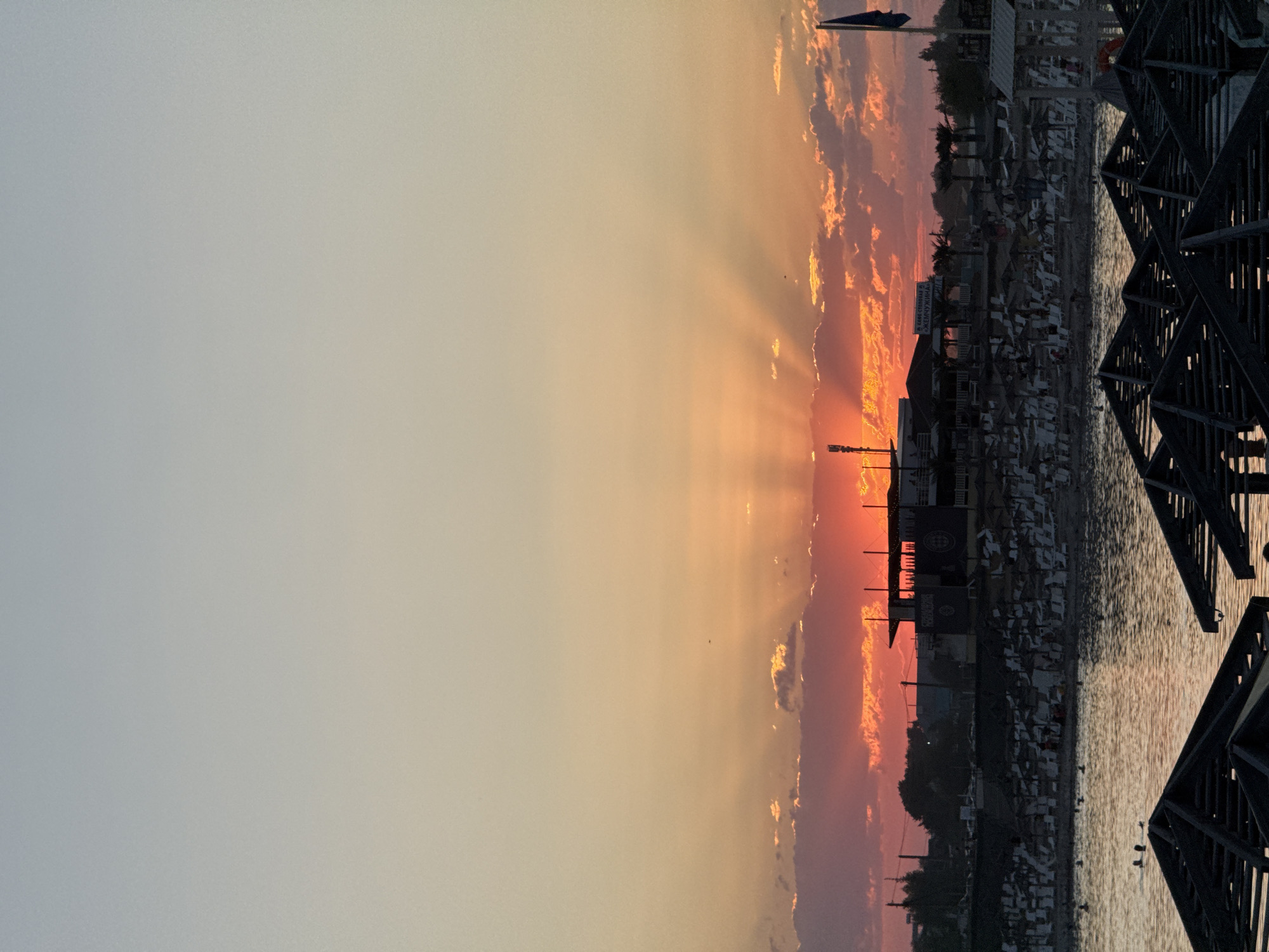 This is a photograph of a beach scene at sunset in Sol-Iletsk, Russia. The sun is low on the horizon, casting rays of light through the clouds and illuminating the sky with vibrant orange and pink hues. In the foreground, several empty beach umbrellas are visible, their dark structures contrasting with the colorful sky. Beyond the umbrellas, a body of water reflects the sunset. On the shore, numerous beach chairs and umbrellas are arranged, with a few people visible in the water swimming. Buildings and palm trees line the beachfront, and a tall tower or antenna is situated on the left side of the frame. A flag flies from a pole on the right side of the image. The overall impression is one of a tranquil evening at a popular coastal resort.