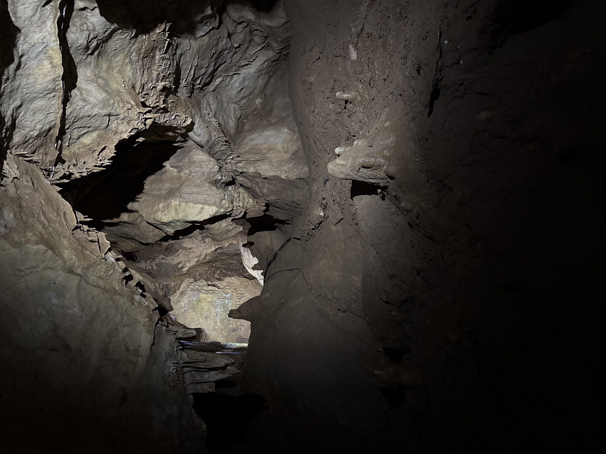 This is a dimly lit cave in Tskaltubo, Georgia. The walls are composed of rough, layered rock formations, with varying shades of brown and gray. The cave narrows into a passage further ahead, illuminated by a faint light source. The foreground is dominated by shadows, creating a stark contrast with the slightly brighter area at the end of the passage. No people or distinct objects are visible other than the natural rock formations.