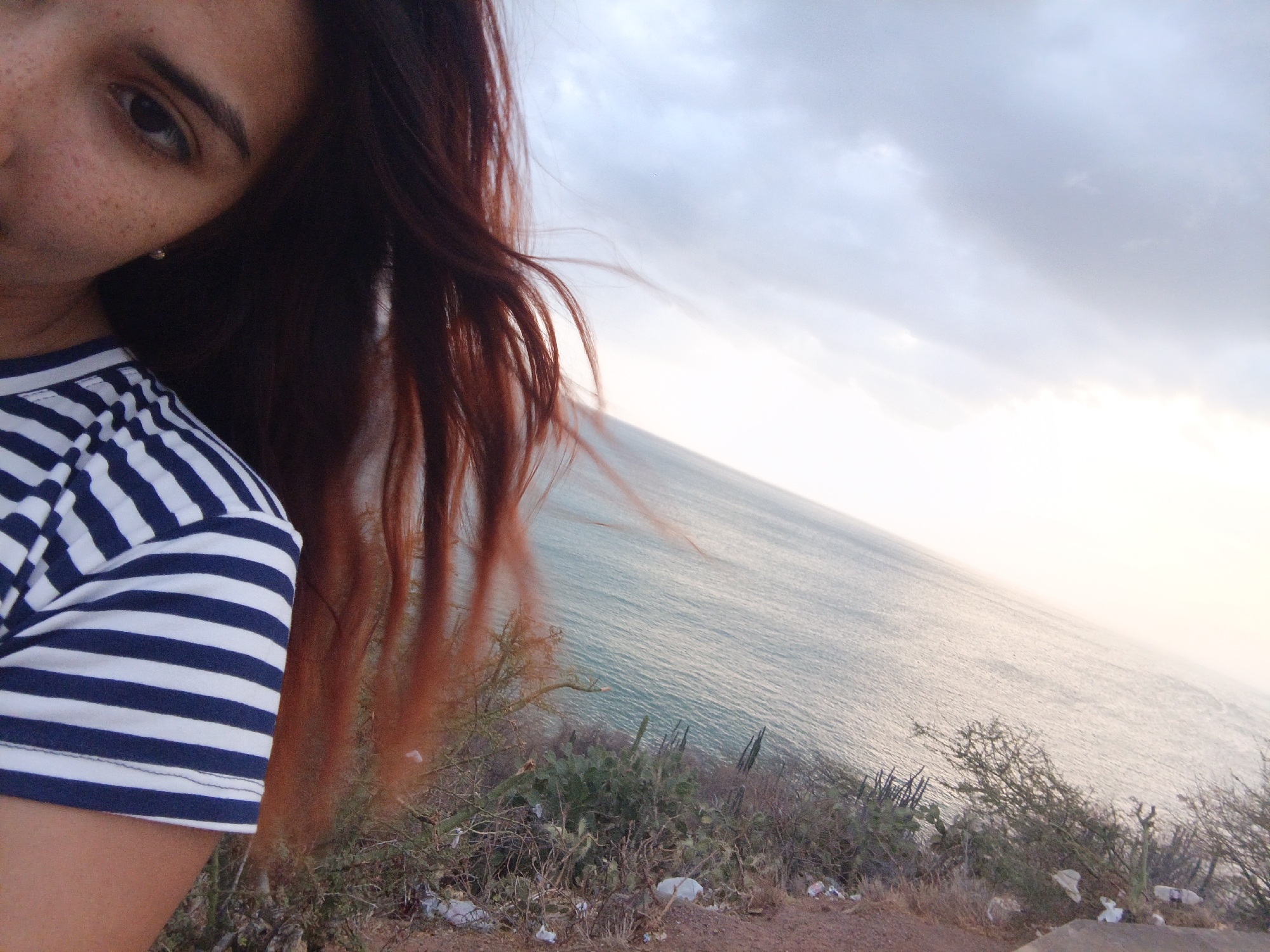 A selfie of a woman with auburn hair, wearing a navy and white striped shirt, taken from a high vantage point in Juan Griego, Venezuela. The scene includes the ocean in the background under an overcast sky and vegetation on the edge of a cliff. The woman's face is partially visible on the left, showing an eye and freckled skin.