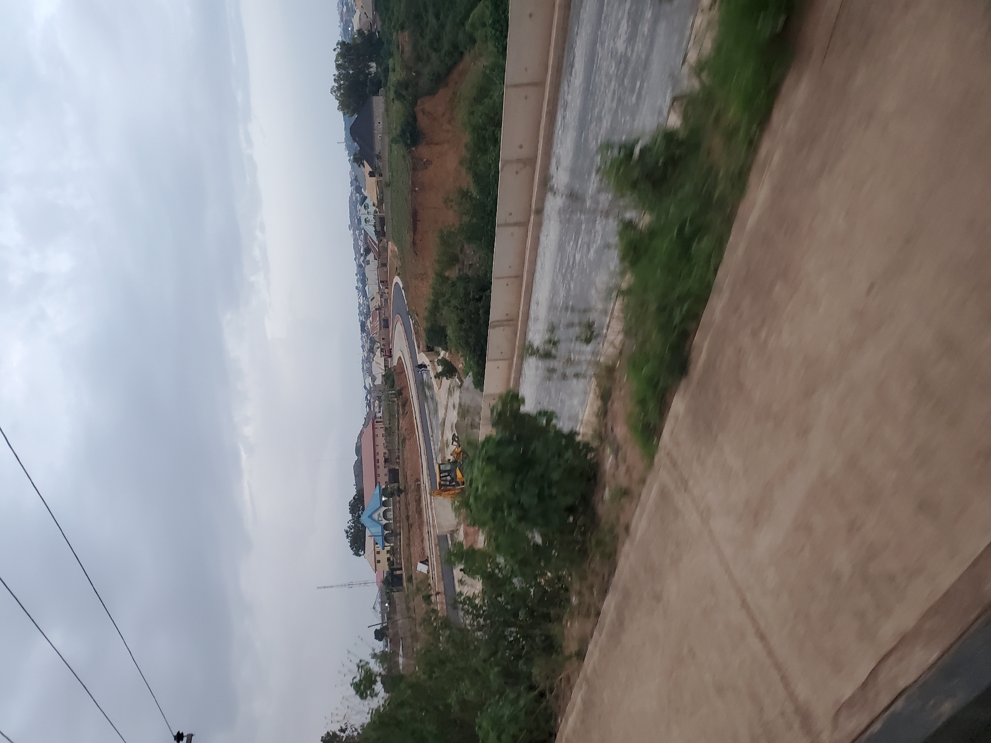 The image captures a view of ongoing construction in Gura Top, Nigeria. In the foreground, a concrete pathway, possibly part of a road or walkway, runs diagonally across the frame. Beyond this, a multi-lane road under construction is visible, with a yellow bulldozer positioned on the left side. The road leads towards a cluster of residential buildings in the middle ground. These buildings exhibit varied architectural styles, with some featuring pitched roofs and others flatter designs. The landscape is characterized by gentle slopes and patches of vegetation, with some areas appearing to be cleared for further development. In the background, a more expansive view of a town or city unfolds, with numerous houses and buildings extending towards the horizon. The sky is overcast with clouds.