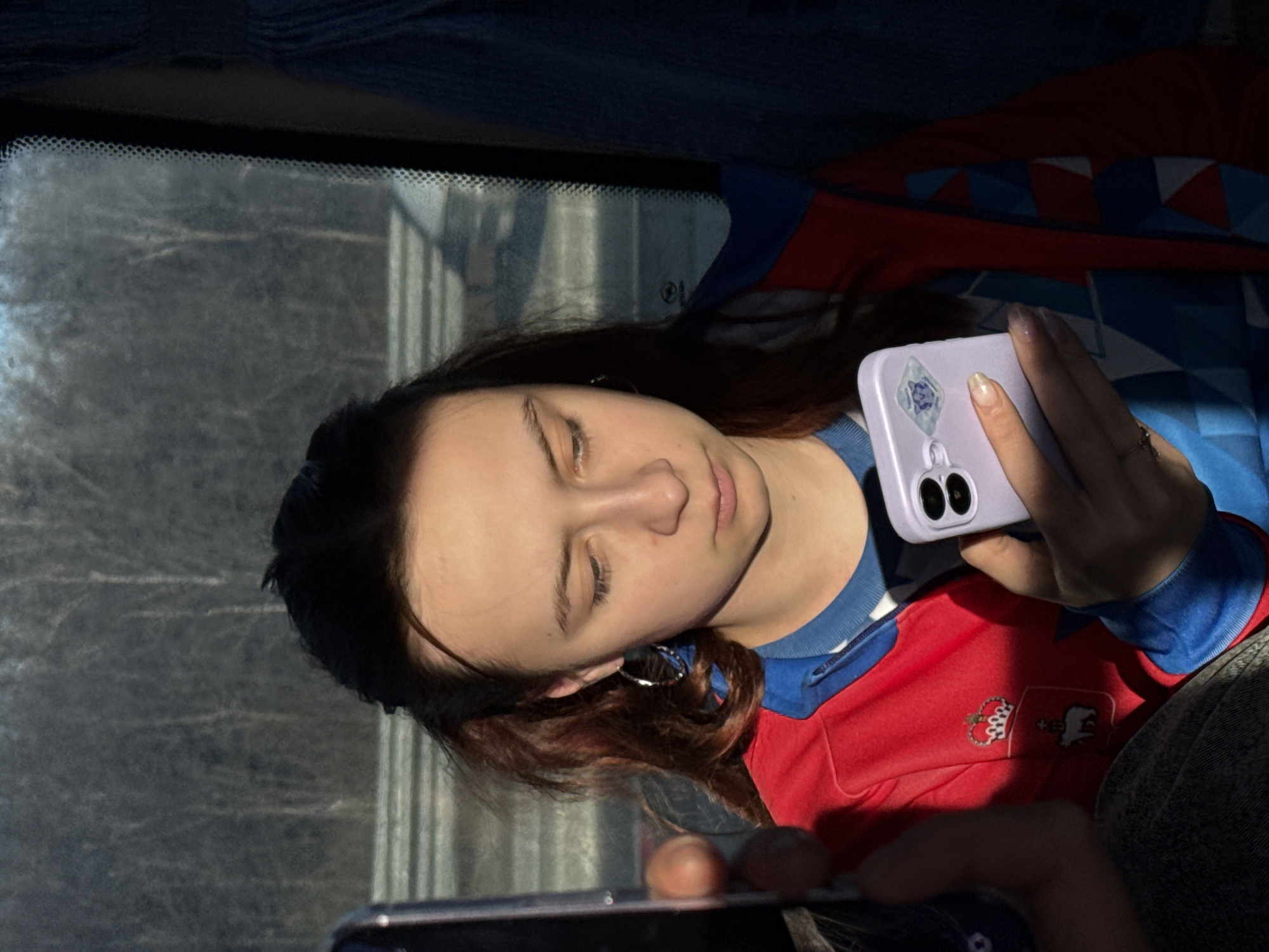 A woman is looking at a phone while sitting in a vehicle, possibly a bus. The woman has dark hair and is wearing a red and blue shirt. She is holding a light purple phone in her right hand. Another phone is visible in her left hand. The background shows a window, suggesting she is inside a vehicle. The location and city are not discernible from the visual information provided.