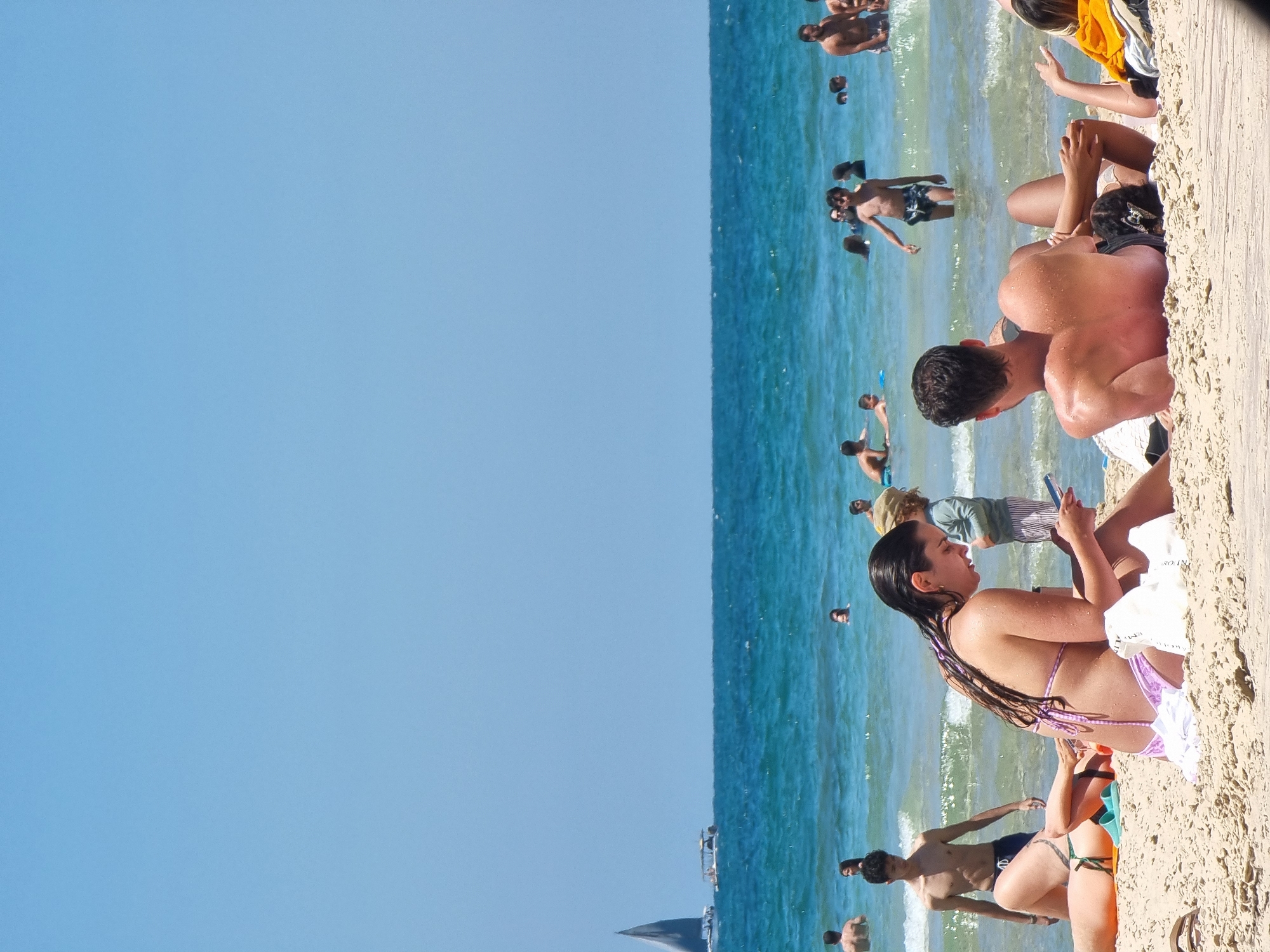 The image captures a vibrant scene at a beach in Tel Aviv, Israel, characterized by clear blue skies meeting the turquoise waters of the Mediterranean Sea. Numerous individuals are present, some actively engaged in swimming and playing in the water, while others are lounging on the sandy shore. A woman with long, dark hair, seated on the sand in a bikini, is looking at her phone. There are others near her on the beach. The overall atmosphere is summery and relaxed. A sailboat is visible on the horizon.