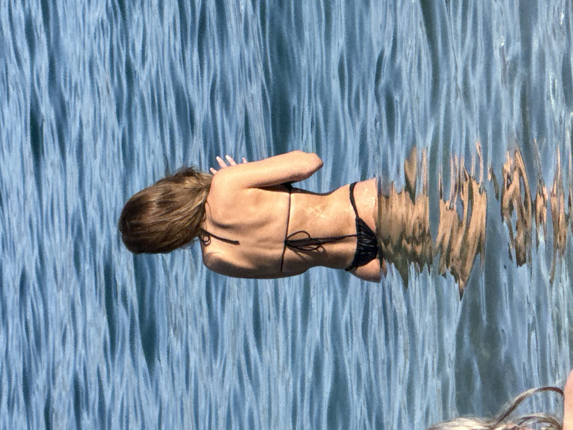 A medium shot shows a woman from behind, standing in the ocean in Budva, Montenegro. She is wearing a black bikini and her hands are raised, palms facing outward. The water is a deep blue and appears to be choppy. The woman's hair is brown and falls to her shoulders. Her skin is tanned. The lighting suggests it is daytime, with the sun reflecting off the water.