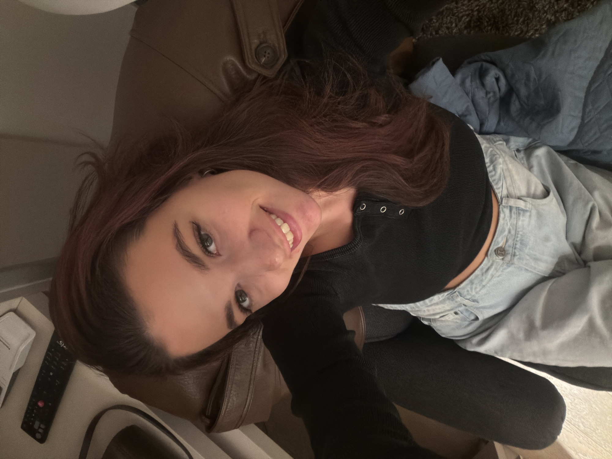 An indoor, high-angle photograph depicts a young adult female seated in a brown leather-like armchair, smiling directly at the camera. She has long, wavy brown hair with reddish highlights and is wearing a black, long-sleeved, ribbed crop top with three light-colored buttons on the placket, along with light blue, faded denim jeans. Her left leg is clad in dark leggings or tights, and a dark blue fabric is draped over her lower body. To her upper left, a black remote control with red and white buttons and a white object are positioned on a light-colored surface. The flooring transitions from a light, possibly tiled or wood surface on the left to a dark, shaggy carpet on the right. The specific location within the city of Belgrade, Serbia, cannot be determined from the visual information presented.