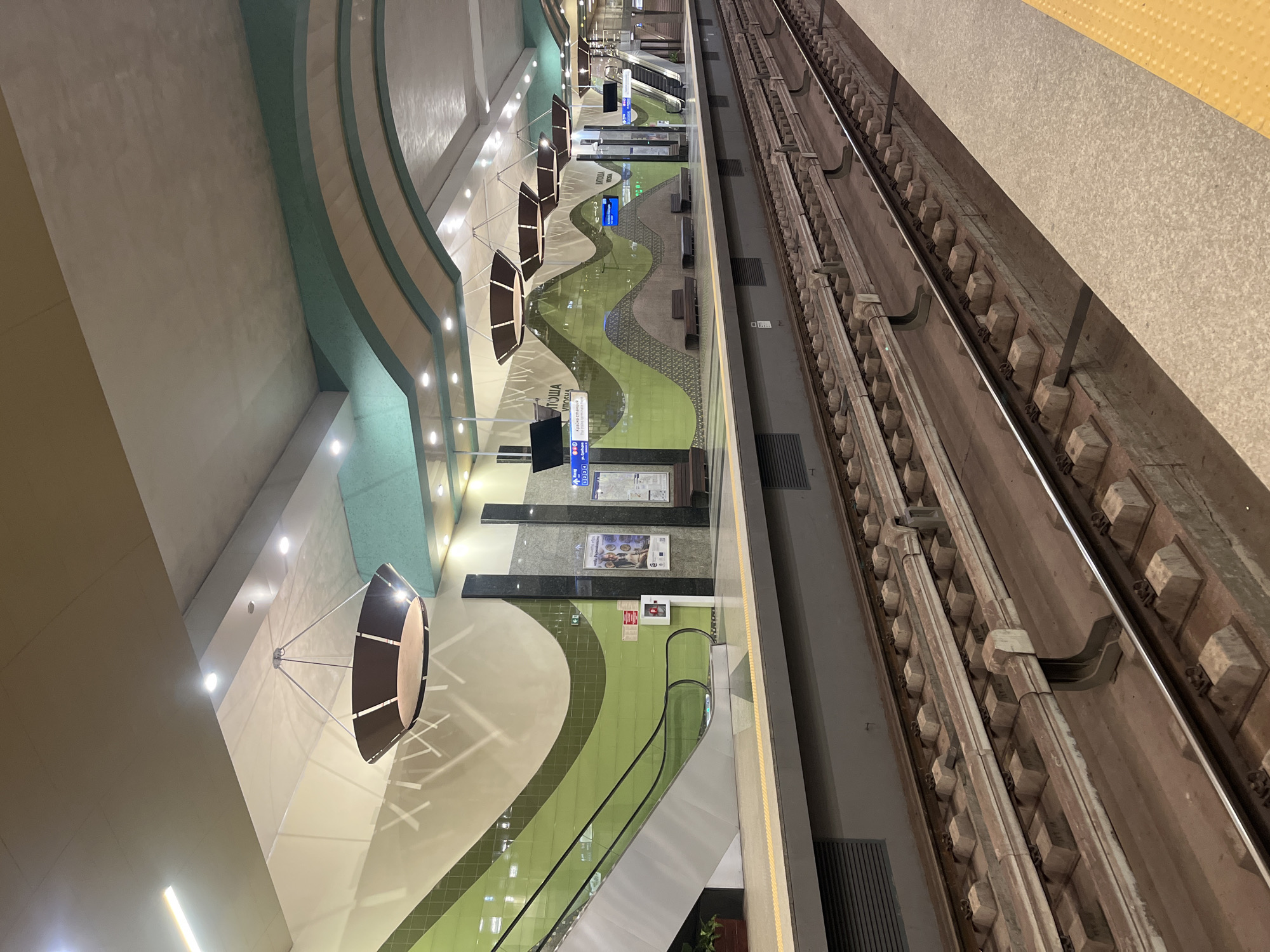 This media file captures an empty, modern metro station platform and tracks in Sofia, Bulgaria, from an elevated perspective, likely during the evening or night given the reliance on artificial lighting. No people are visible.
On the right side of the image, two sets of metro tracks stretch into the distance, constructed with robust concrete sleepers and metal rails. A narrow platform edge, marked with a yellow textured warning strip, runs parallel to the tracks.
The metro platform, located on the left, showcases a distinctive and contemporary design. The floor features undulating patterns in light green and cream/beige, complemented by darker grey sections. The adjacent walls are adorned with curved architectural elements in light blue-green and beige tones.
Several large, drum-shaped light fixtures are suspended from the ceiling, casting illuminated patterns and shadows on the surfaces. Additional recessed spotlights provide general illumination throughout the station. Dark-colored benches are strategically placed along the platform for seating.
Information and directional signage are prominent. Blue overhead signs with white arrows indicate directions. A large sign displays text in Cyrillic, reading "ГОША" (GOSHA) and "КРАЙНА СТАНЦИЯ" (KRAYNA STANTSIYA), which translates to "End Station." Another sign includes "VIKTORIYA" in Latin script. Various posters and advertisements are affixed to the walls.
At the far end of the platform, an escalator is visible, connecting to another level, suggesting multi-tiered station architecture. A red fire extinguisher box is mounted on a pillar. The scene conveys a sense of calm and order within a clean, well-designed public transport environment.