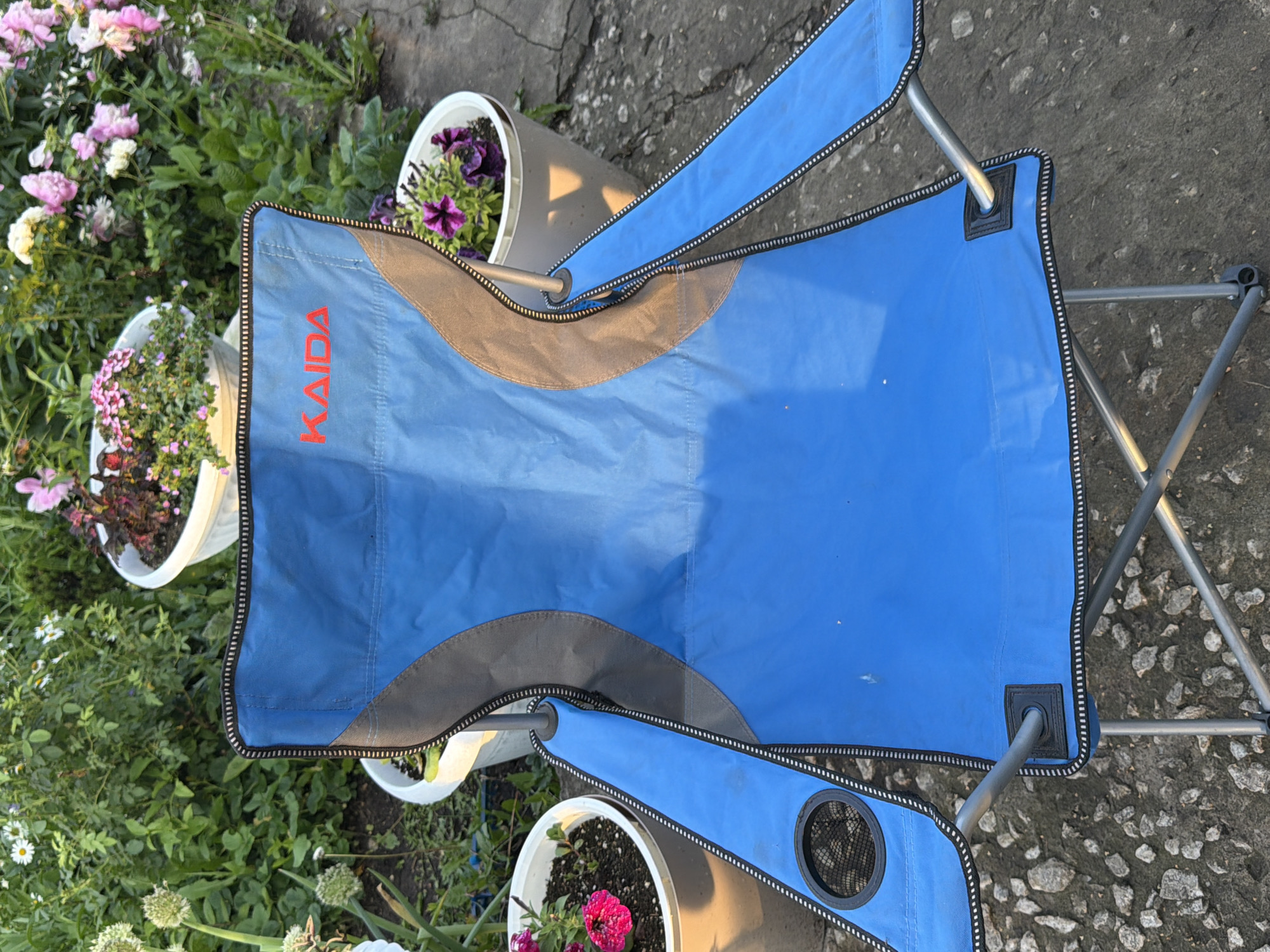 The image shows a blue camping chair with a black and grey trim, featuring the red logo "KAIDA" on the backrest. The chair is placed outdoors on a cracked concrete surface, possibly a patio or sidewalk. In the background, there are several potted plants with green foliage and colorful flowers, including white daisies, pink petunias, and purple petunias. The vegetation suggests a garden setting. The scene appears to be in Sukhodol, Russia, given the context provided. There are no people visible in the image. The lighting suggests it is daytime, with sunlight casting shadows.