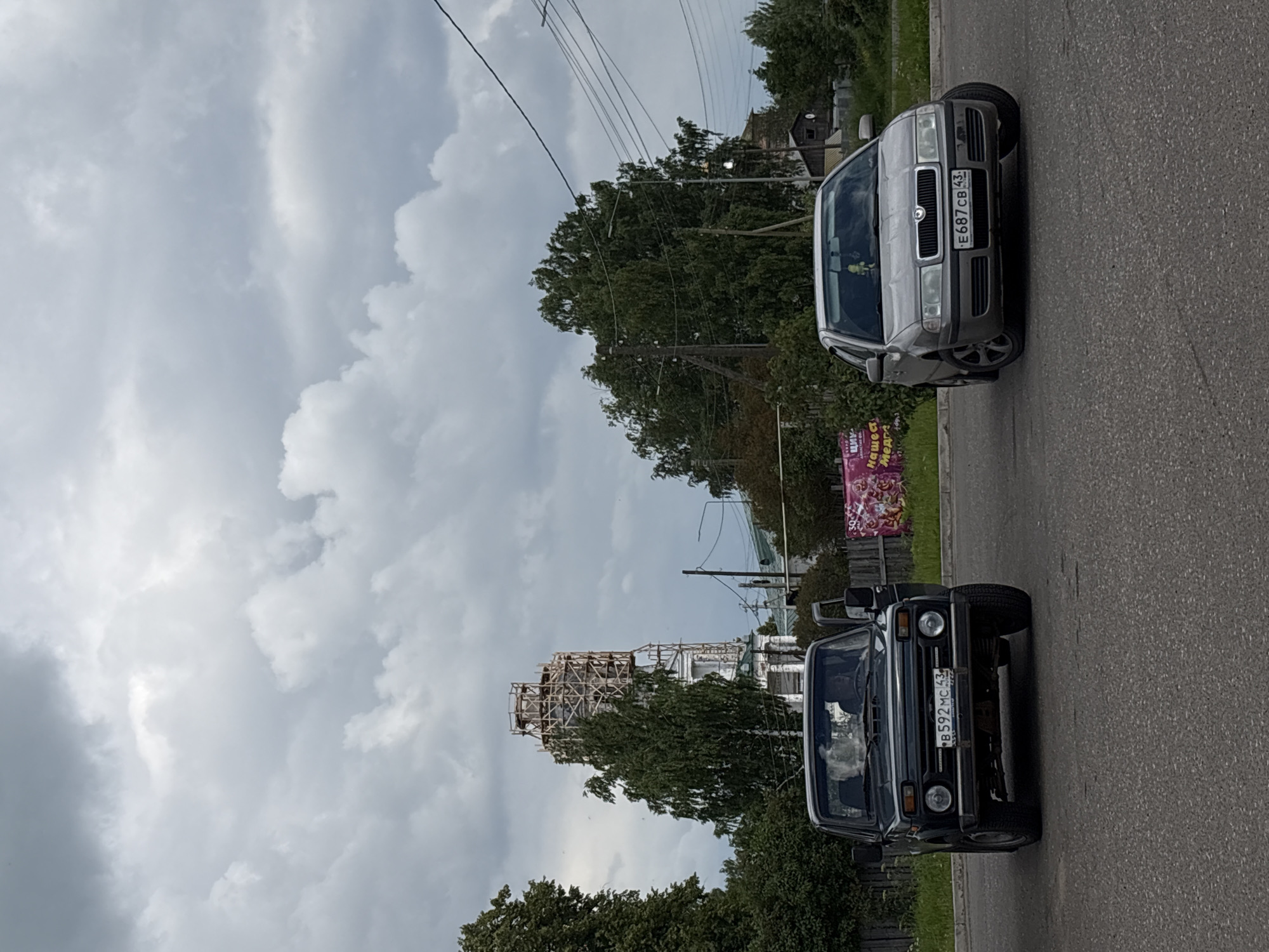 In Кстинино, Russia, under a cloudy sky, two cars are parked on an asphalt road. A dark colored SUV, with its driver's side door open, is on the left. A silver sedan is on the right, its front wheels turned sharply to the left. Behind the cars, a tall, white tower under construction, surrounded by wooden scaffolding, is visible amongst green trees and other buildings. The road also has power lines overhead.