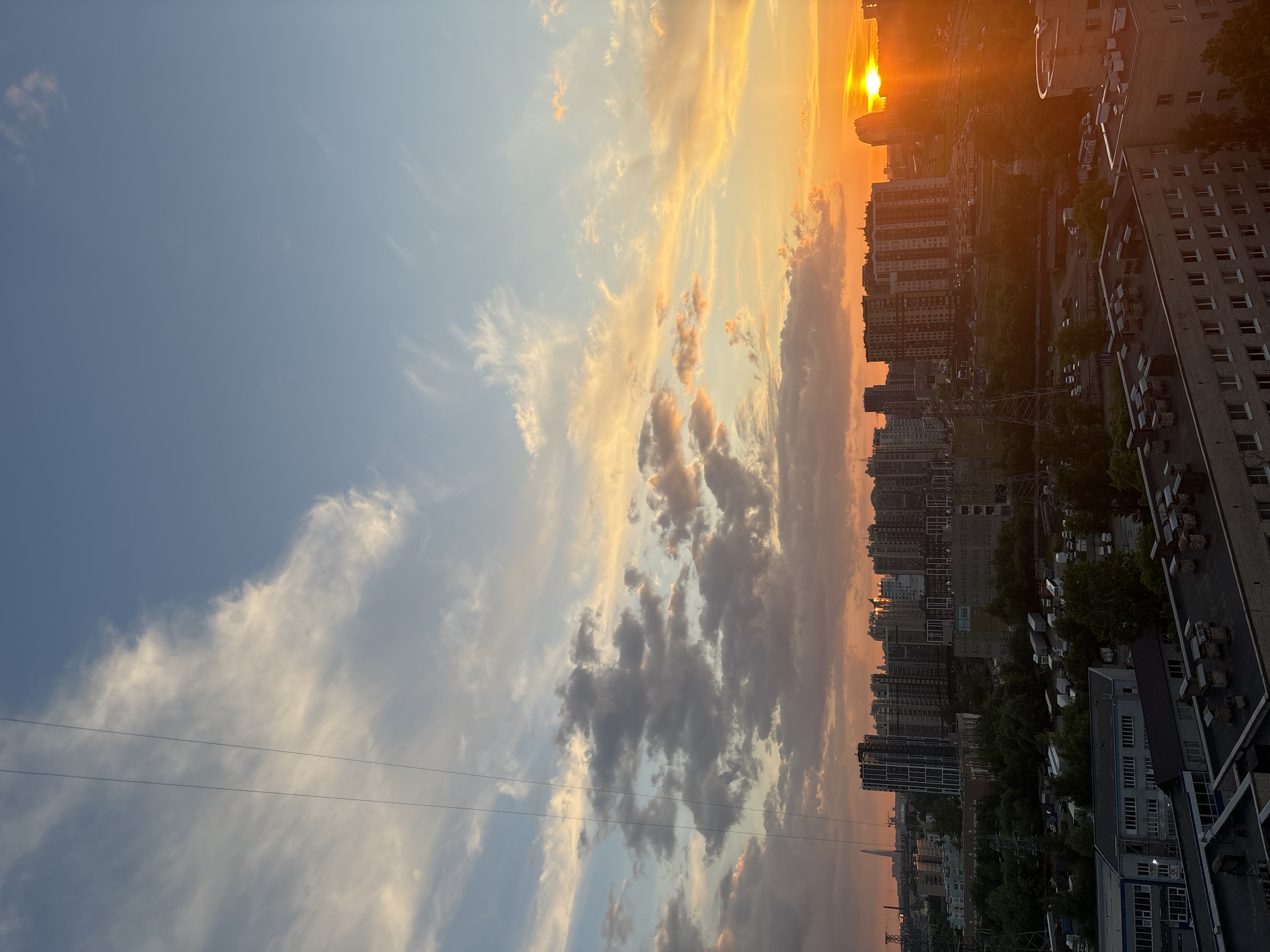 The image displays an urban landscape at sunset or sunrise, viewed from an elevated perspective. The scene is dominated by a vibrant sky transitioning from a soft blue on the left to intense oranges and yellows towards the right, where the sun is visible as a bright, low-hanging orb.
Various types of clouds are scattered across the sky: delicate, wispy formations in the bluer areas, and more substantial, textured clouds tinted golden and pink by the sun's light in the central and right portions. Several thin, dark utility lines stretch horizontally across the middle-left part of the sky.
Below the colorful horizon lies a dense cityscape. Numerous multi-story buildings, likely residential or commercial, form a sprawling silhouette against the bright sky, particularly those closer to the sun. In the foreground and middle ground, some structures are more detailed, revealing windows, rooftops, and equipment. Patches of green trees are interspersed among the buildings, and faint outlines of parked vehicles can be seen in various spots.
The time of day is clearly sunset (or possibly sunrise) due to the low position and intense coloration of the sun and sky. The weather appears clear with scattered clouds, and there's a slight atmospheric haze near the horizon. The scene evokes a sense of calm and beauty typically associated with dusk over a city. No specific human activity is taking place, and there is no visible text in the image. The architectural style and density suggest a contemporary urban environment that aligns with the "Unknown, Russia" location context.