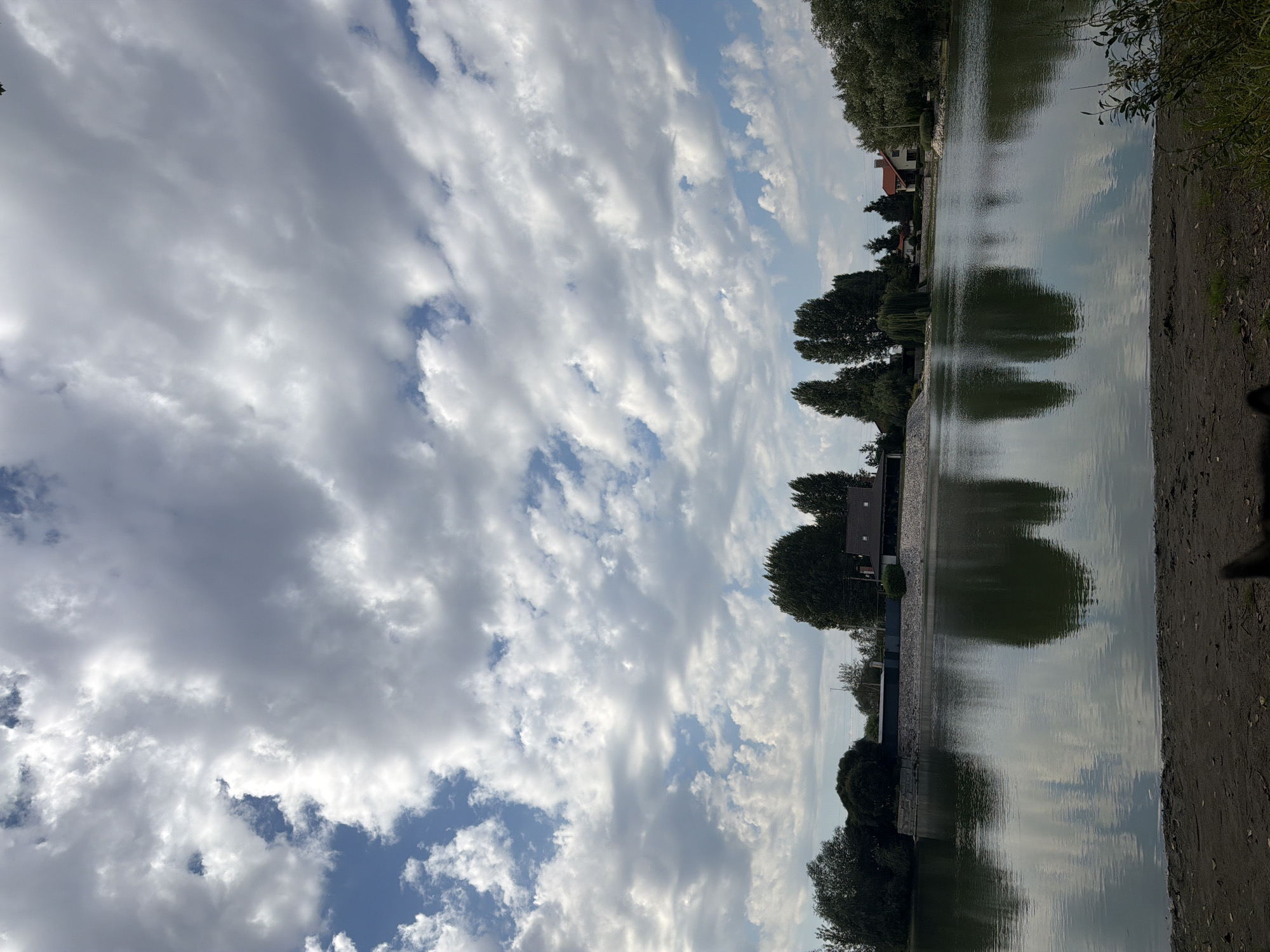 The image shows a serene lake with calm, reflective water. The shoreline in the foreground is composed of dark sand and some sparse vegetation. Across the lake, a lush green bank is lined with mature trees, and a few houses with red roofs are visible in the distance. The sky above is a dynamic mix of blue patches and dramatic, billowing white and grey clouds, suggesting a breezy day. The overall scene is peaceful and natural, with a touch of human settlement visible along the far bank. There is no specific indication of the city of Узюково, Russia, within the image itself.