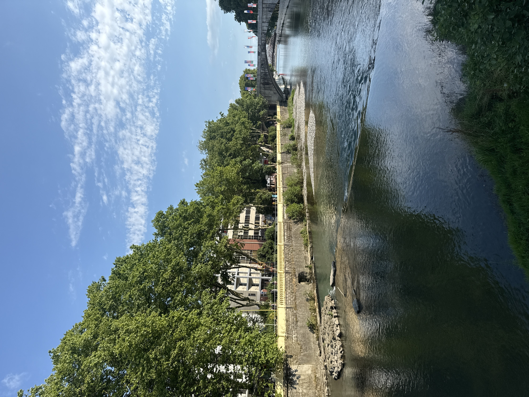 The image depicts a scenic river flowing through an urban environment, likely in Sochi, Russia, on a bright, sunny day with scattered white clouds. The river, with its clear, rippling water, takes up a significant portion of the frame, reflecting the sky and surrounding greenery. The riverbank is lined with lush green trees, their leaves vibrant in the sunlight, casting shadows on the path that runs alongside.
A notable feature is the yellow railing that separates the walkway from the river, providing a clear visual demarcation. Beyond the trees and railing, several multi-story buildings are visible, their architecture suggesting a mix of modern and older styles. The presence of flags on a bridge in the background adds a touch of international flair and hints at a public event or celebration. While no specific activity is clearly depicted, the scene evokes a sense of peaceful leisure, perhaps a stroll along the river or a moment of quiet reflection. The overall atmosphere is tranquil and inviting, typical of a pleasant summer day in a European city. There is no visible text in the image.