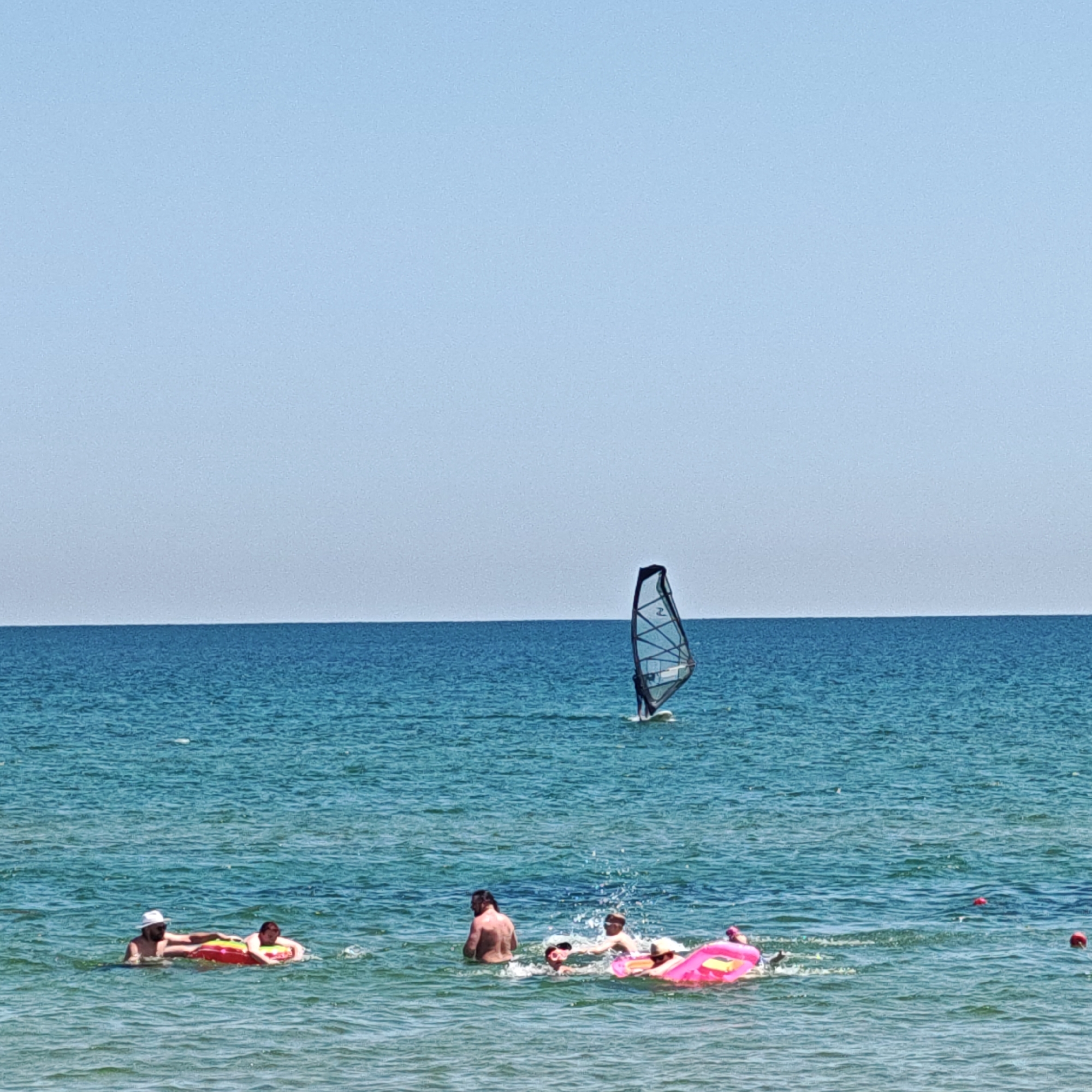 The image depicts a scene at a beach in Мысовское сельское поселение, Ukraine. The vast expanse of the Black Sea occupies the majority of the frame, with a clear, light blue sky above. The water is a vibrant turquoise, with gentle waves lapping the shore.
In the foreground, several people are enjoying the water. On the left, two individuals are floating on a large, circular inflatable raft. One person, wearing a white hat, is propped up on the raft, while the other, a child, is partially submerged and leaning on the raft. Further to the right, a group of people are interacting in the water. One person, a man with dark hair, is standing in the water. Next to him, a child is on a bright pink inflatable raft, and another person appears to be seated on the same raft.
In the mid-ground, a lone windsurfer is visible, skillfully maneuvering their board and sail across the water. The sail is dark with white accents, and the surfer is positioned on a white board. The windsurfer is a moderate distance from the shore, suggesting an active day on the water.
The overall impression is one of a serene and sunny day at the beach, with people engaging in various recreational activities. The clear water and bright sky suggest pleasant weather conditions.