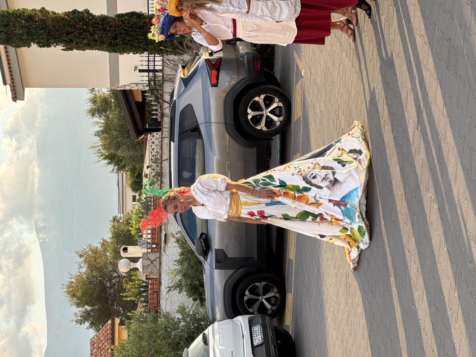 In Isola delle Femmine, Italy, a woman stands on a paved surface, looking to her right. She is wearing a white, long-sleeved shirt with puffed sleeves and a voluminous, white skirt adorned with colorful illustrations of fruit, faces, and roosters. A yellow sash cinches her waist. She has red and green feathers in her hair. To her right, two other women are partially visible, also wearing similar white tops and red skirts, one with a blue hat decorated with flowers and the other with a yellow hat. Behind them, a gray SUV is parked, and a white Dacia Sandero is parked to the left, with its rear visible. The background shows residential buildings, trees, and a hazy mountain range under a cloudy sky. The ground is paved with interlocking gray bricks, and long shadows are cast across it by an unseen object to the left.