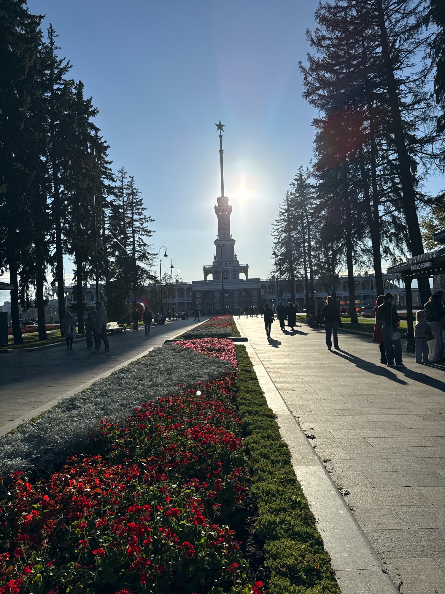 A wide, paved walkway is lined with flowerbeds filled with red and white flowers, and manicured green hedges. Tall evergreen trees flank the walkway, casting long shadows. In the background, a large, ornate building with a central tower topped by a star is visible. The sun is shining brightly, creating lens flare and silhouetting many of the people walking on the path. Several individuals are seen strolling along the walkway, some in small groups and others alone. The overall scene suggests a public park or plaza on a sunny day.