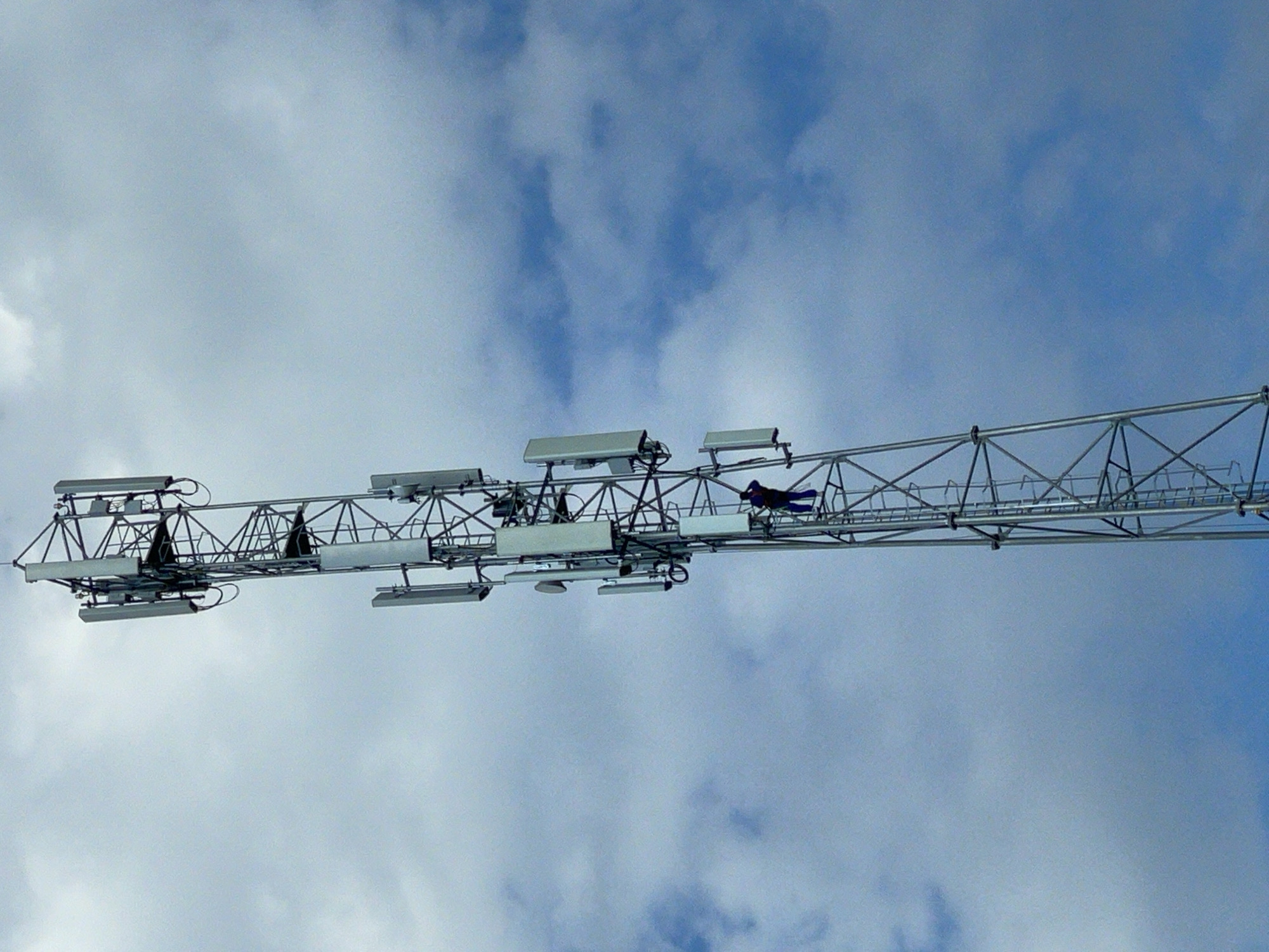 A telecommunications tower stands tall against a sky with scattered clouds. The tower is constructed of metal lattice work and is equipped with multiple antennas. A lone individual, wearing a dark blue outfit and a safety harness, is positioned on a ladder-like section of the tower, appearing to be performing maintenance or an inspection. The scene suggests an industrial or urban setting, with the sky indicating daytime conditions. The tower is a dominant feature, with the person on it being a small but significant detail, highlighting the scale and operational nature of the structure. The location is identified as Piskivka, Ukraine.