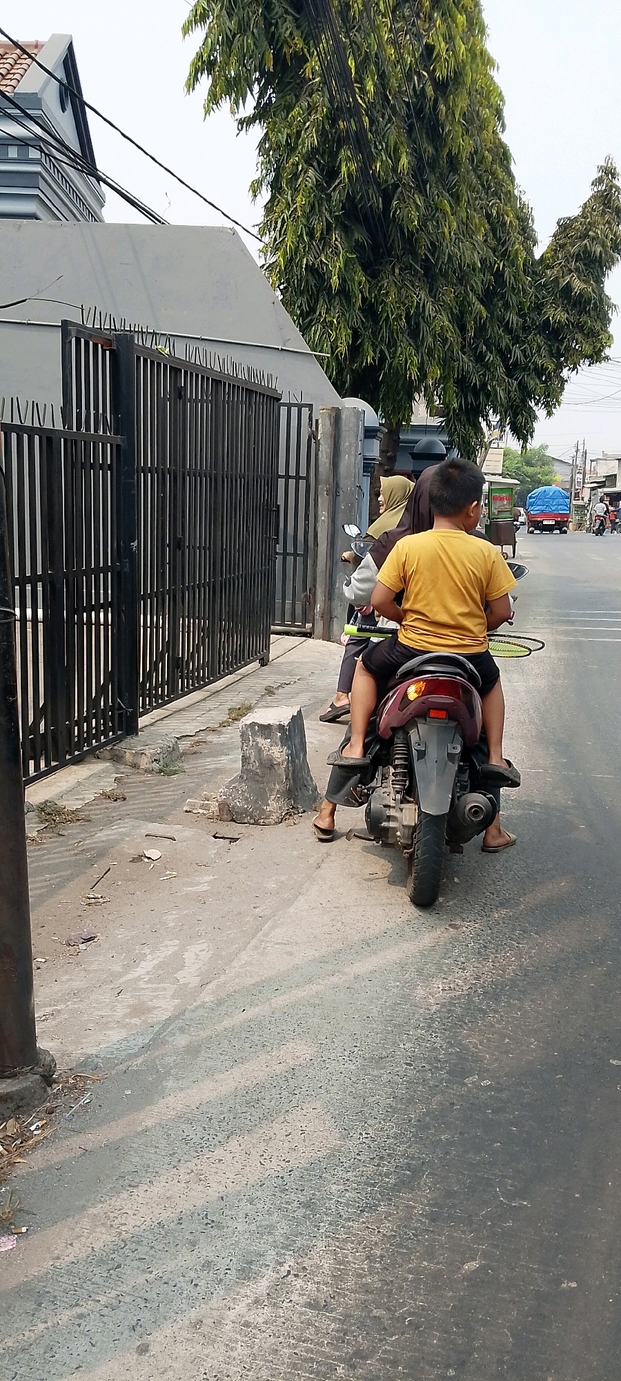 The image depicts a street scene in Bekasi, Indonesia, on a bright, overcast day. In the foreground, a child wearing a yellow t-shirt and dark shorts sits behind an adult on a maroon scooter. The child holds a green badminton racket horizontally in front of them. The adult, wearing a hijab and grey long-sleeved shirt, is also on the scooter. The scooter is positioned on the right side of a paved road.
To the left of the scooter, a black metal fence borders a property with a grey-toned building visible beyond it. The fence has vertical bars and a large gate. In front of the fence, there is a sidewalk made of grey concrete. A broken concrete block sits on the edge of the sidewalk, and debris, including leaves and what appears to be cigarette butts, is scattered on the ground.
Further down the road, other vehicles are visible, including a blue truck with a covered bed. The environment appears to be urban or suburban, with trees and buildings lining the street. The overall impression is that of a typical day in a bustling Indonesian city.