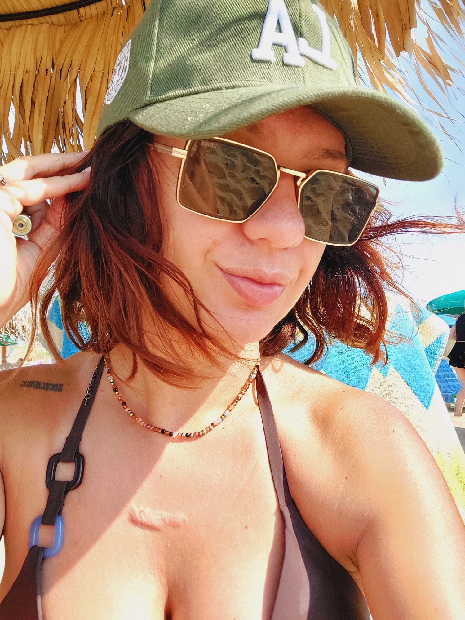 A medium shot captures a woman from the chest up, outdoors under a thatched umbrella. She wears a green baseball cap with a white emblem and sunglasses with gold frames. Her short, auburn hair frames her face. She is also wearing a brown bikini top and a beaded necklace. A scar is visible on her chest. To her left, a Roman numeral tattoo is visible on her shoulder. The background includes a blue and yellow striped towel and a beach umbrella. The image appears to be taken on a sunny day at a beach.
