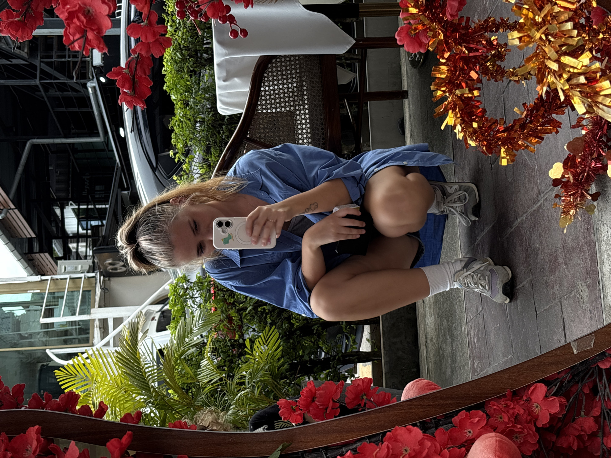 A young woman is reflected in a mirror, taking a selfie with her phone. She is crouched down, wearing a blue shirt and white socks with sneakers. She has blonde hair pulled back, and a tattoo is visible on her left arm. The mirror reflects an outdoor scene with lush green plants, red flowers, and a glimpse of a white vehicle and building structures. Decorative red and gold tinsel hangs in the upper right portion of the reflection, suggesting a festive atmosphere or season. The lighting appears to be natural daylight. The setting seems to be a public space, possibly a cafe or restaurant patio, given the presence of chairs and tables reflected in the background. The overall mood is casual and relaxed. The text "llowh" is visible, mirrored, on a white structure in the background reflection.