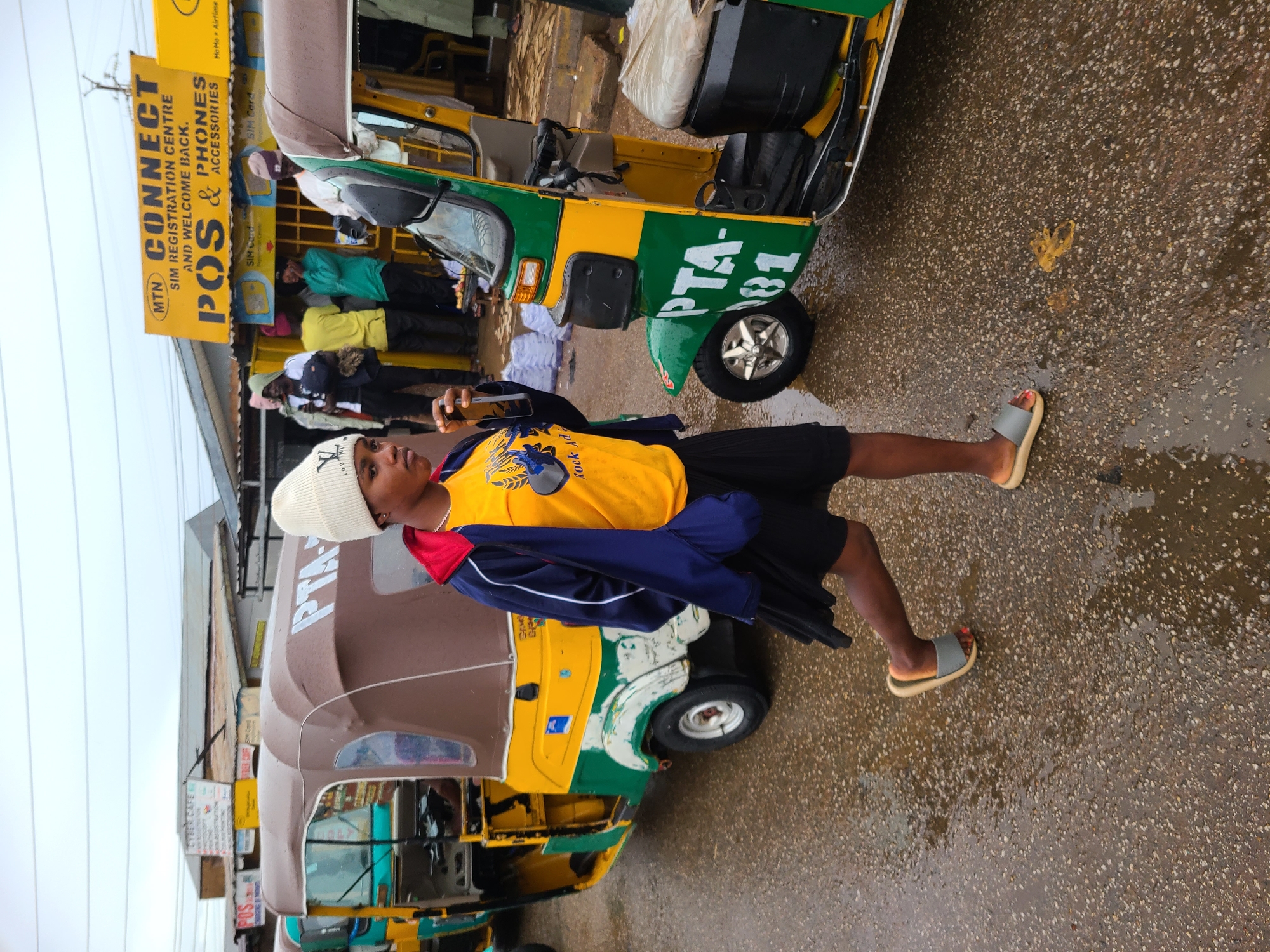 A young man stands in front of a row of auto-rickshaws on a wet street in Gura Top, Nigeria. He wears a white beanie, a yellow t-shirt, a blue jacket, and black shorts, and sandals with a red stripe on the toes, and he holds a cell phone in his hand. Behind him is a brown auto-rickshaw and behind that is a yellow and green auto-rickshaw that has “PTA-81” on the side. A yellow sign with the words “MTN CONNECT SIM REGISTRATION CENTRE AND WELCOME BACK. POS & PHONES & ACCESSORIES” is visible behind the rickshaw. Other people are standing in the background under the sign.