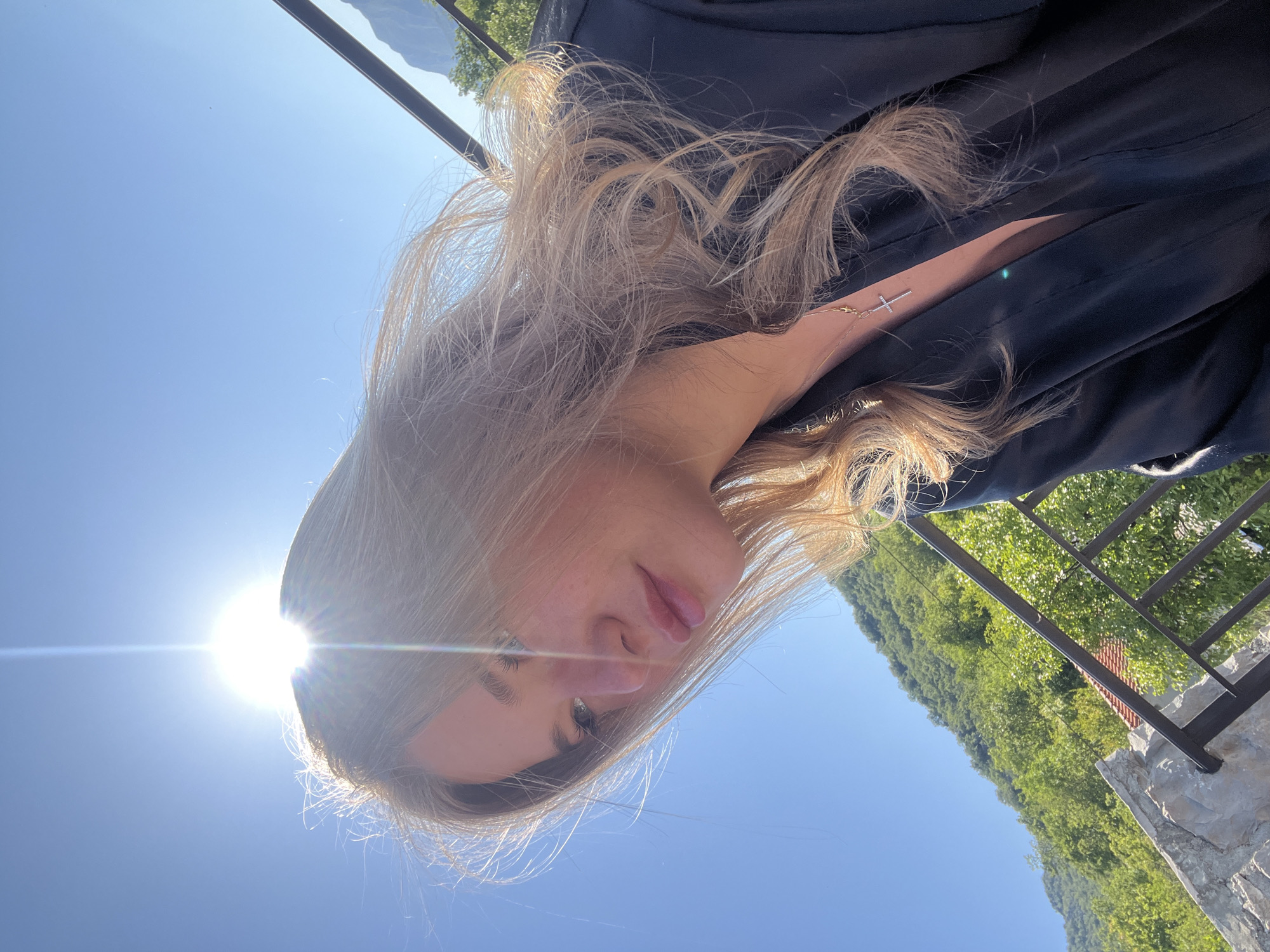 A woman is standing outside in Sotonići, Montenegro. The sun is shining brightly, and the sky is a clear blue. She is wearing a dark jacket and a necklace with a cross pendant. The background shows a hillside covered in green trees and a stone railing.