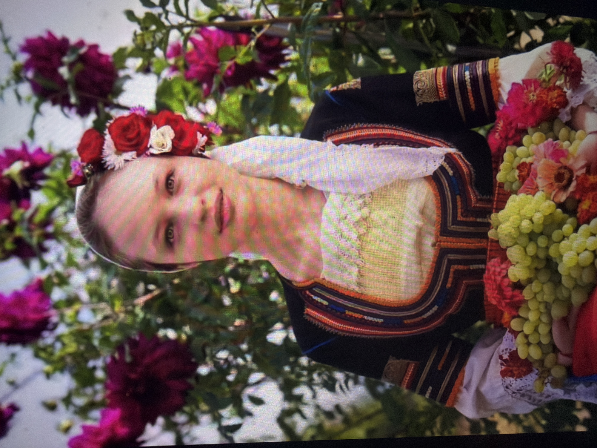 The image displays a young woman with fair skin and light eyes, positioned centrally in a vertical composition within a garden setting in Shumen, Bulgaria. Her hair is styled back, partially concealed by a white headscarf that drapes over her left shoulder. A floral arrangement, consisting primarily of red roses, smaller white flowers, and subtle pink accents, is attached to the right side of her head. She is dressed in traditional Bulgarian attire, which includes a long-sleeved black outer garment featuring intricate multi-colored embroidery in patterns of red, orange, blue, gold, and brown on the chest, shoulders, and cuffs. A white blouse with lace detailing is visible beneath the outer garment at the neckline and sleeve ends. The woman is holding a substantial quantity of light green grapes and several pink and red flowers in her arms. The background consists of dense green foliage interspersed with large, vibrant purple and magenta flowers.