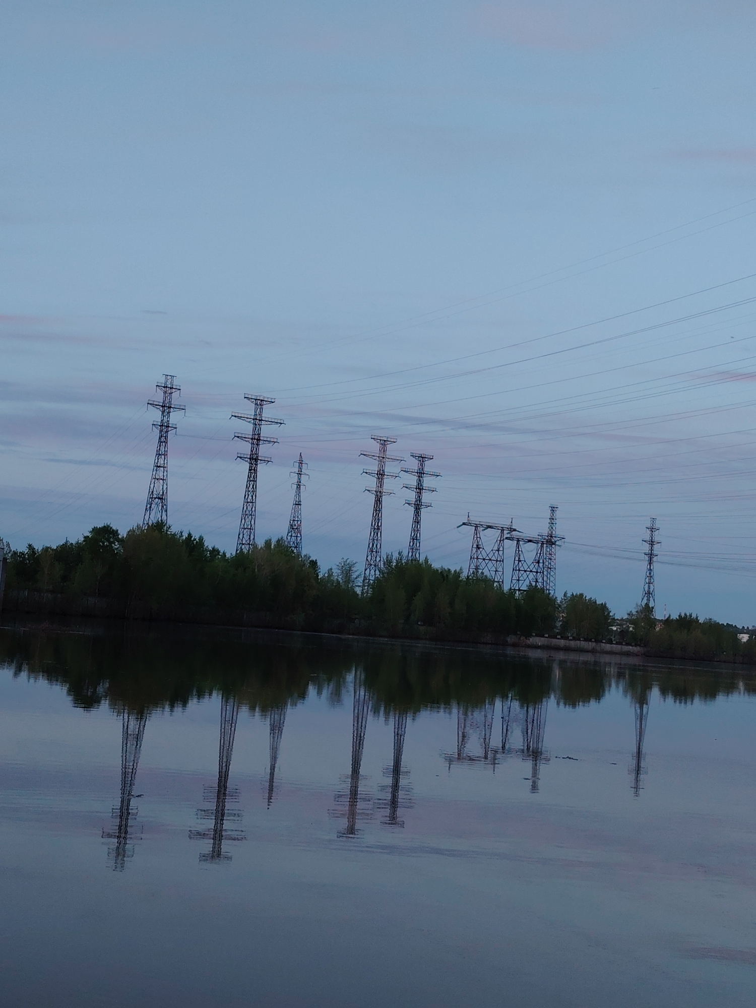 The image depicts a serene twilight scene at the Kama River in Chaikovsky, Russia. Several large electrical transmission towers are situated along the riverbank, their forms reflected in the water. Lush green vegetation borders the shoreline. High-voltage power lines extend from the towers across the sky. The sky displays a gradient of pale blues and subtle pink hues. The water surface exhibits a slightly rippled texture, mirroring the silhouettes of the towers and trees.