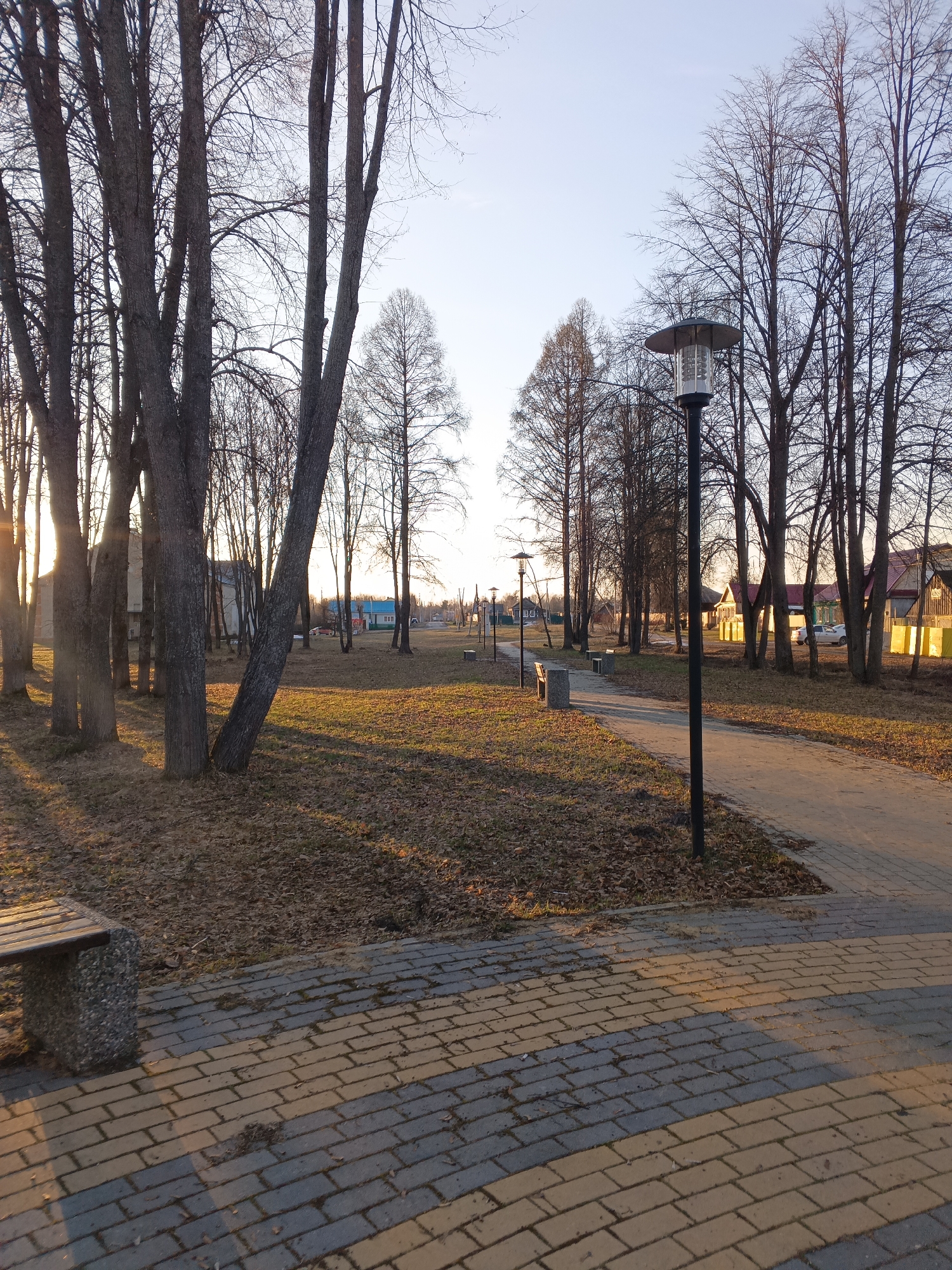 The image shows a park in Красный Маяк, Russia. A paved walkway runs through the center of the park, lined with bare trees and lampposts. A bench is visible in the foreground. The ground is covered with dry grass and fallen leaves. Some buildings are visible in the background. The lighting suggests it is either sunrise or sunset. There are no people visible in the scene.