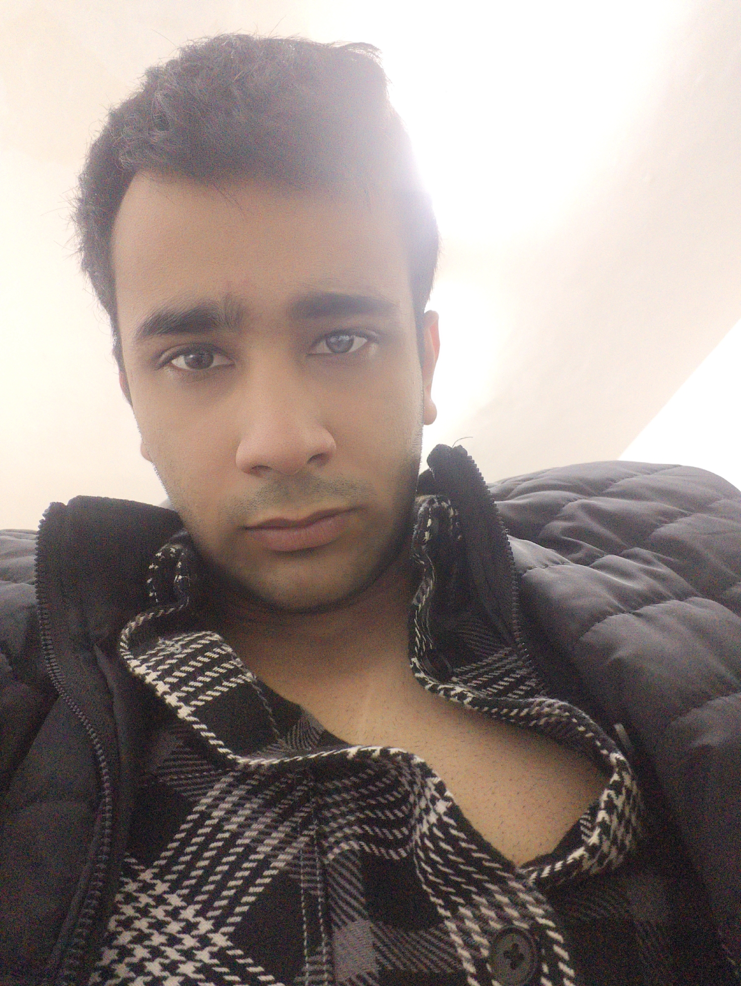 A close-up selfie depicts a young man with dark hair, eyes, and complexion. He's wearing an open black and white patterned shirt beneath a black quilted jacket. The background is a plain, bright, indeterminate color, likely a wall or ceiling. The lighting appears to be artificial and slightly harsh. No indicators point to Chongqing High-tech Zone, China. There's no discernible environmental context provided in the image.