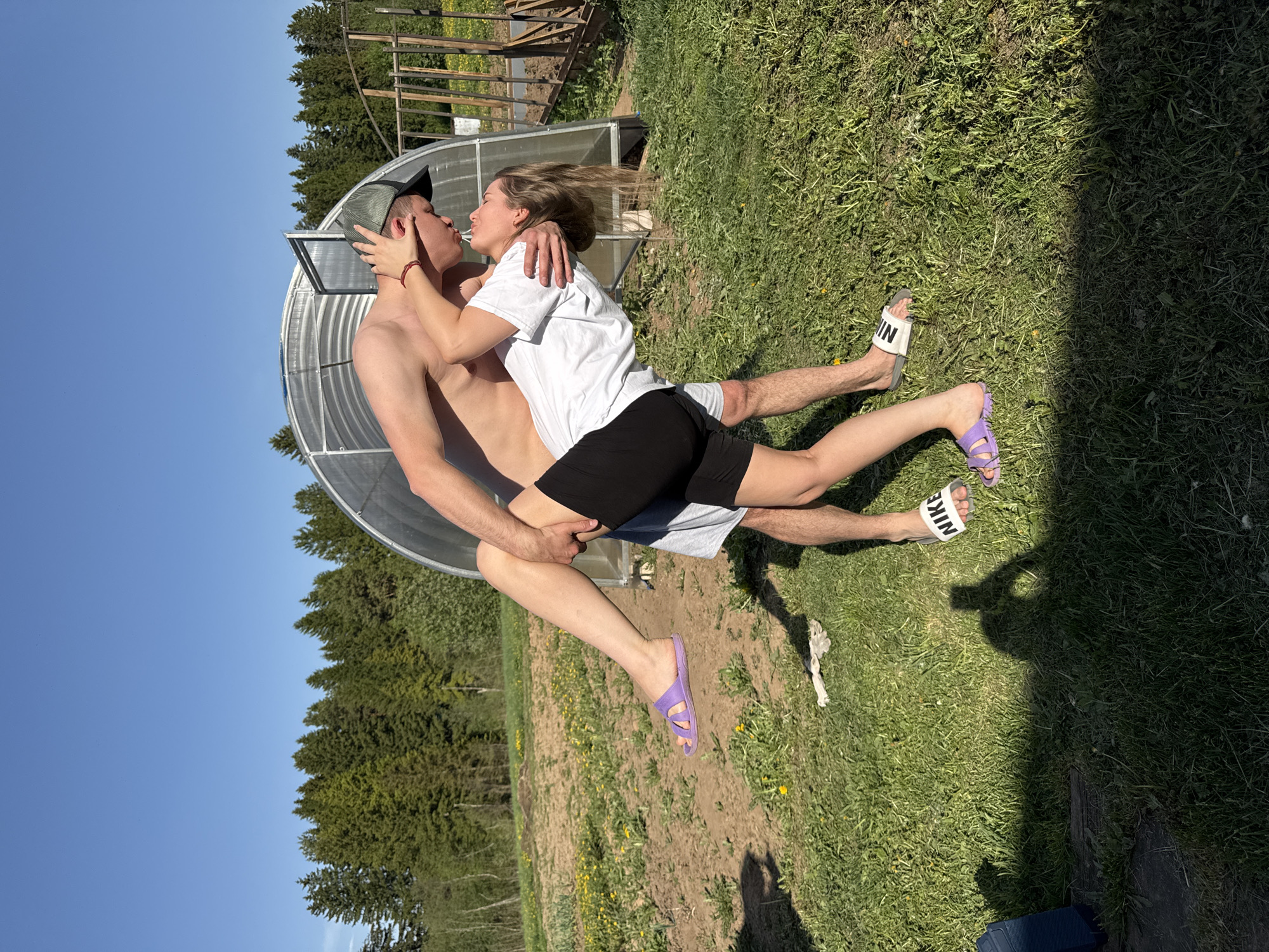 The image shows a couple kissing outdoors in a sunny, grassy area. The man, shirtless and wearing a cap, is holding the woman, who is wearing a white t-shirt and black shorts. The woman is wearing purple sandals and the man is wearing white Nike sandals. Behind them is a greenhouse and trees. The sky is blue. It is daytime. There is no evidence this photo was taken in Фоки, Russia.