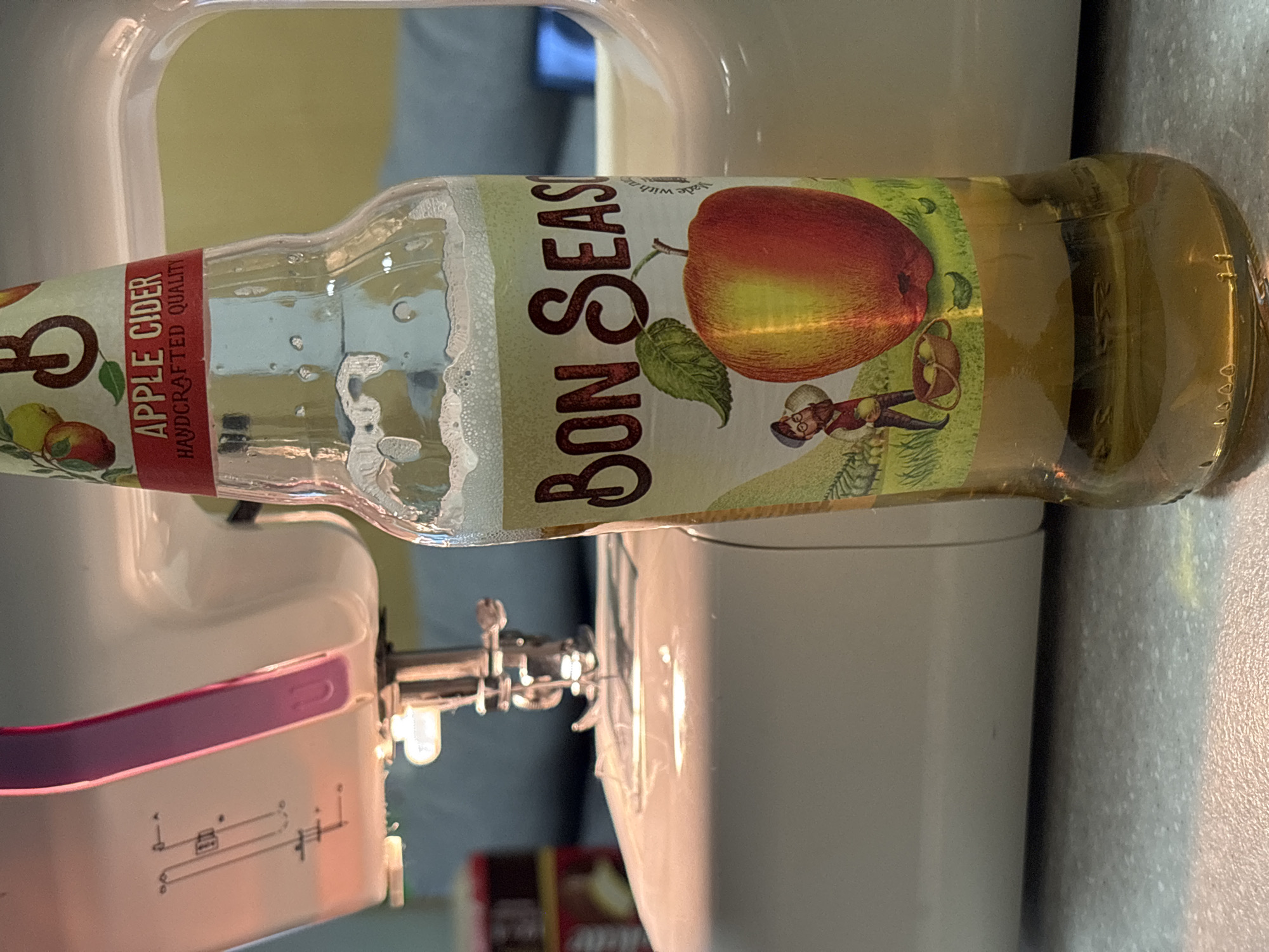 A bottle of Bon Season apple cider rests horizontally atop a white sewing machine. The label is visible, featuring an apple and an illustration of a person carrying a basket. The sewing machine's needle and presser foot are visible on the left. The background includes blurred shapes of furniture and a wall. There is no information to identify the location in the city of Perm, Russia.
