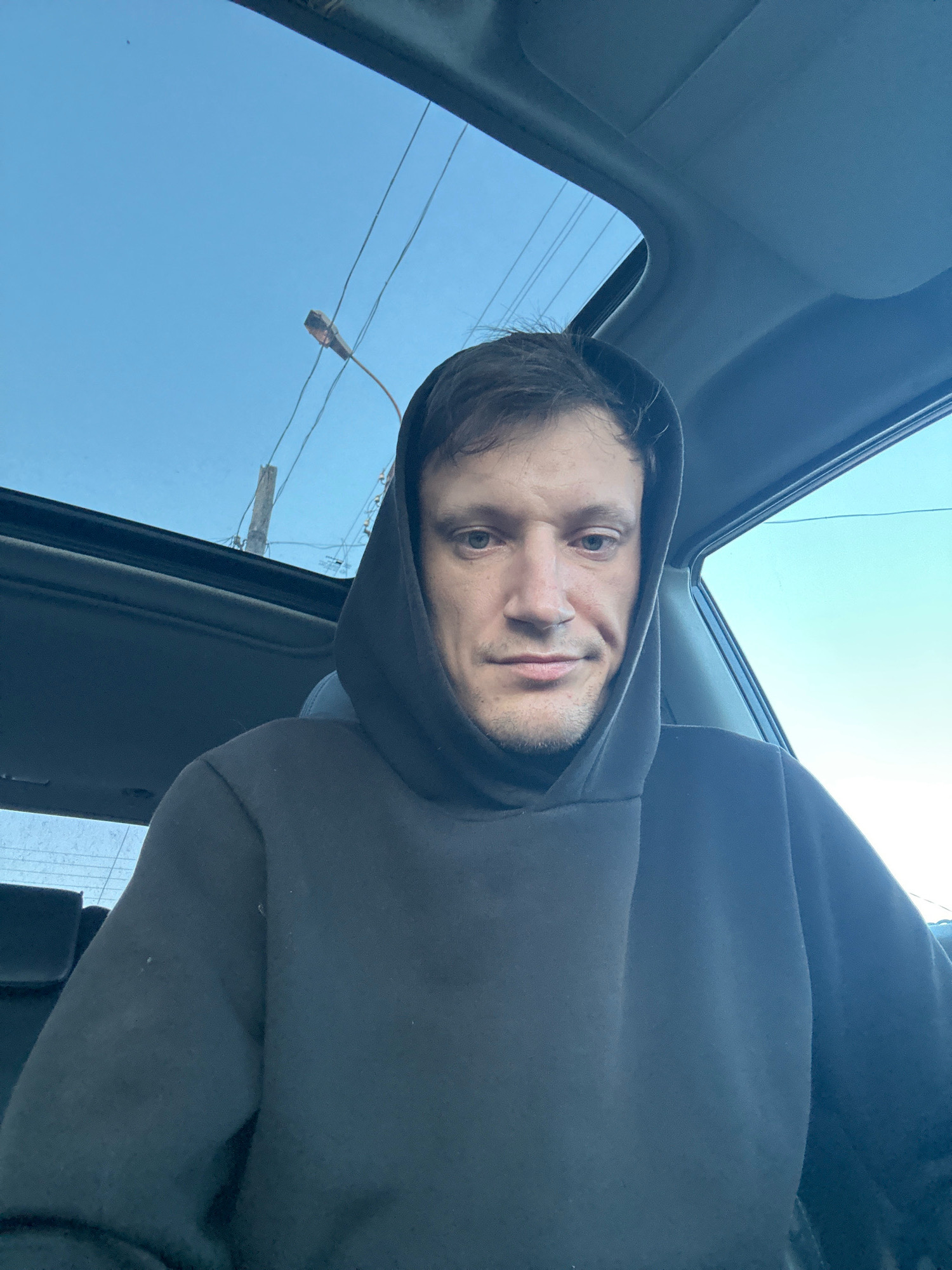 A man wearing a black hooded sweatshirt is sitting in a car. The sunroof of the car is open, revealing a blue sky with power lines and a street lamp visible. The man is looking directly at the camera with a neutral expression. The interior of the car, including a headrest, is visible in the background. The photo appears to be taken during daylight.